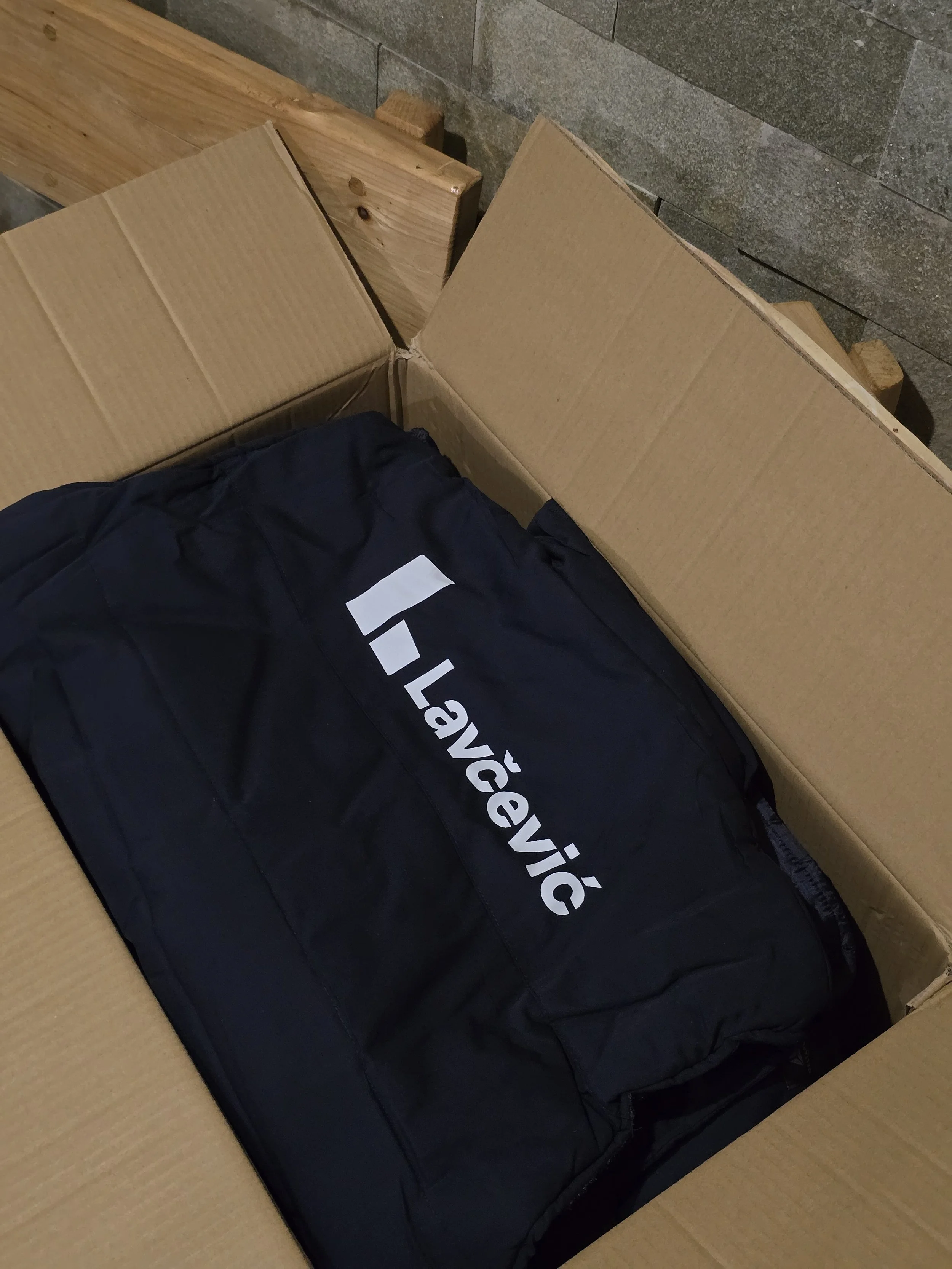 A cardboard box containing a black jacket or bag with white text that appears to spell 'ÉLLEV' or similar, resting on a wooden surface next to a grey stone wall.