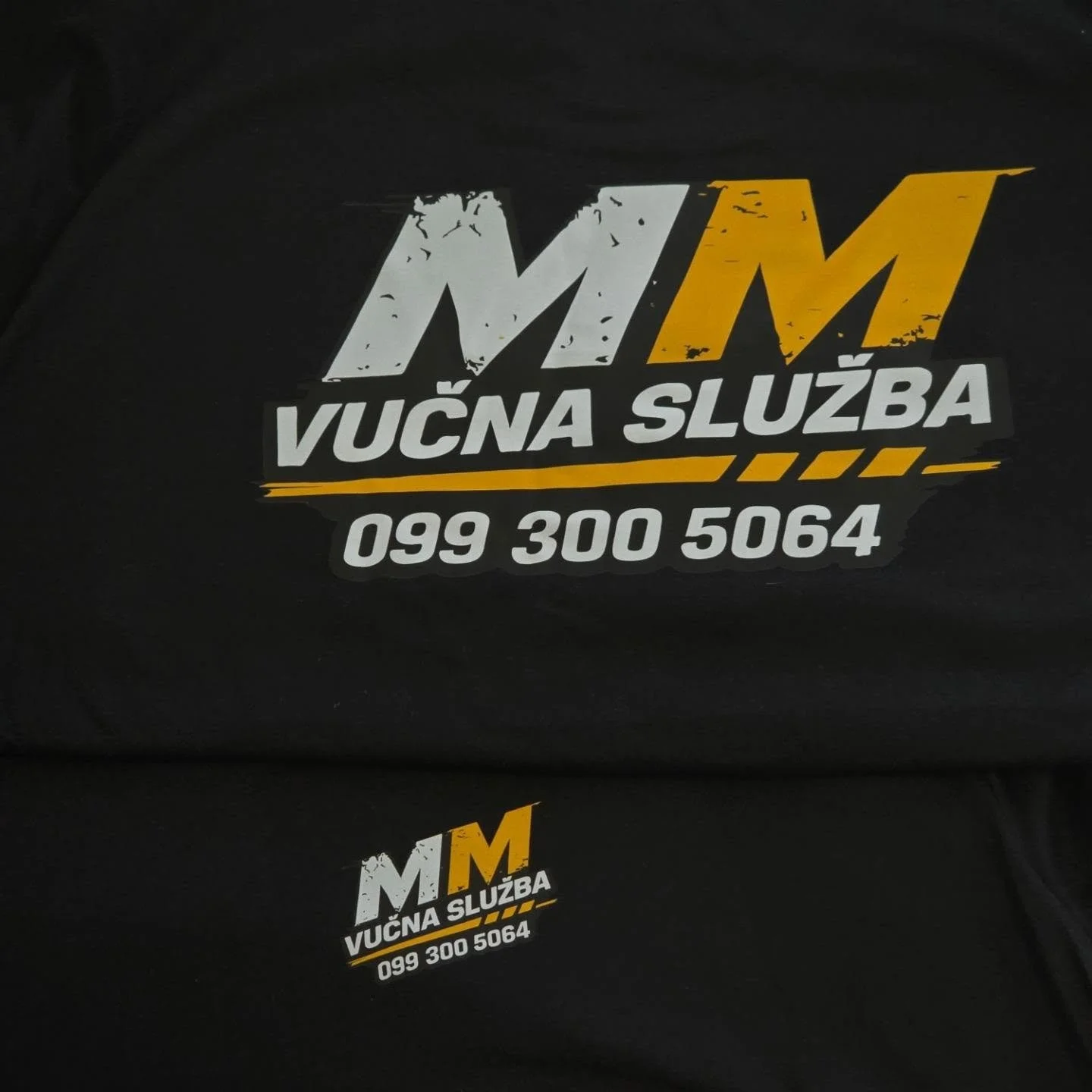 Sticker on a black surface displaying the logo and contact information for 'MM Vučna Služba', a towing service. The sticker has a black, white, yellow, and gray color scheme.