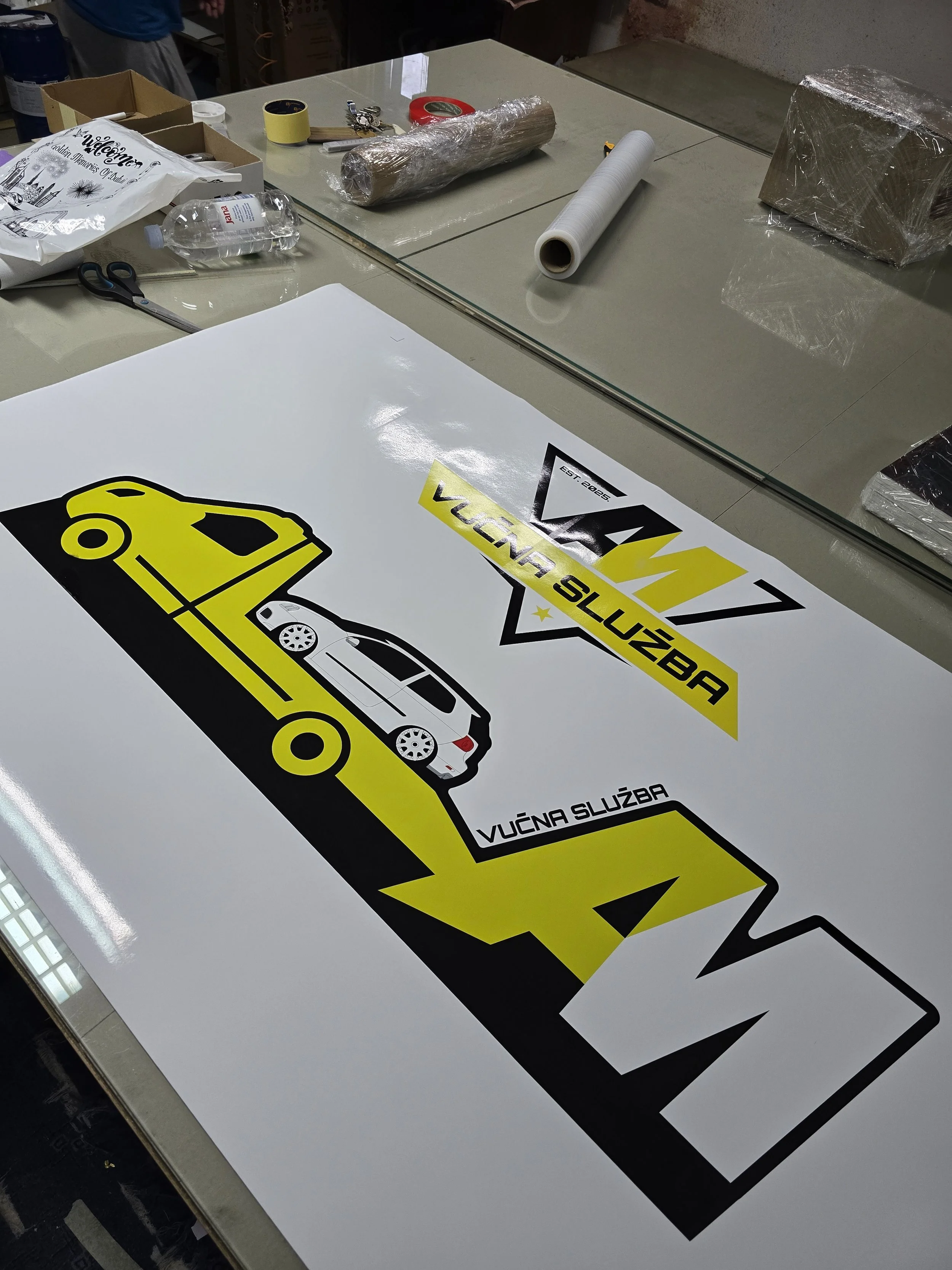 A table with a large sticker that has a black and yellow design of a car and text that says 'VUCNA SLUZBA'. Around the table, there are various items including a water bottle, scissors, tape, rolls of vinyl or adhesive material, and a cardboard box.