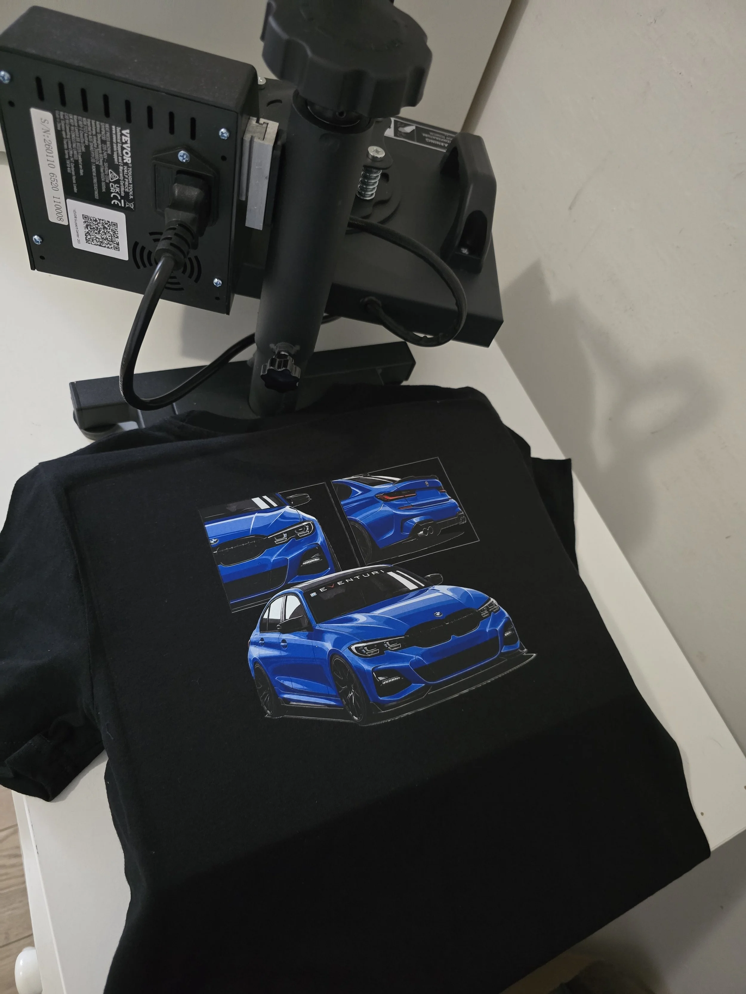 A blue sports car printed on a poster, placed on a black T-shirt, which is on top of a black bag. A corner of a printer is visible in the background.