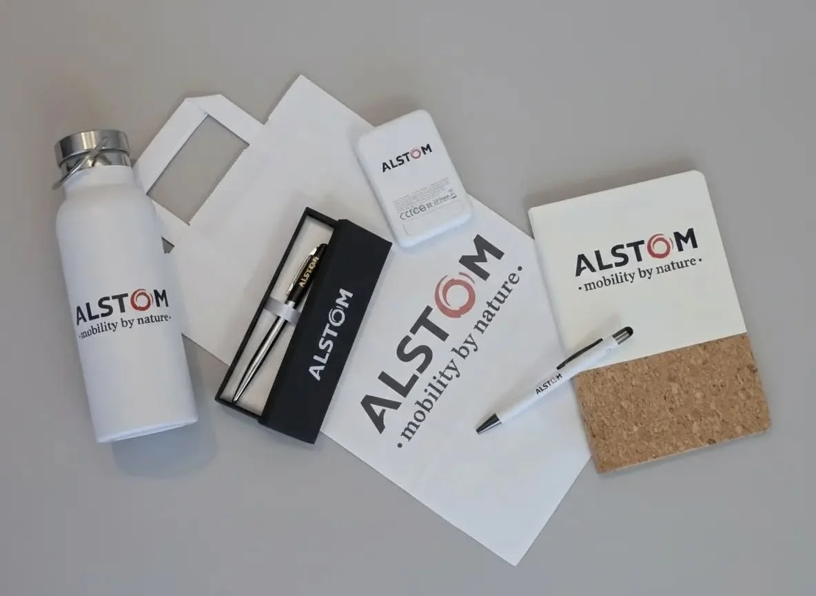 Alstom branded water bottle, white paper bag, white sheets of paper, small white box, black box with pen, cork notepad with Alstom logo, black and white pen with Alstom branding, all items on a gray surface.