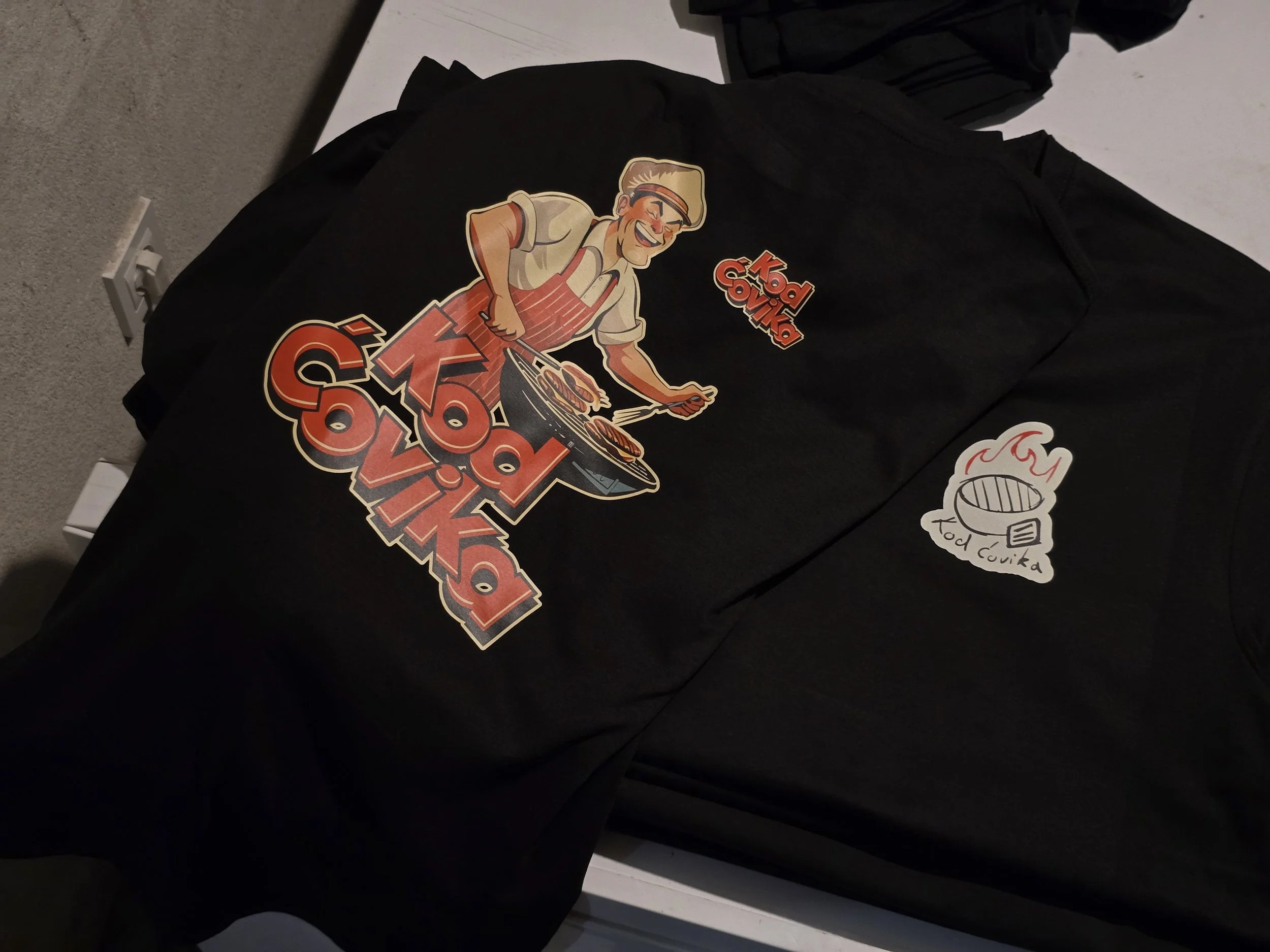 Black t-shirt with a cartoon image of a man with sunglasses, chef's hat, and apron grilling, along with the text 'KBBQ PDX' and a sticker reading 'Food Cueva'.