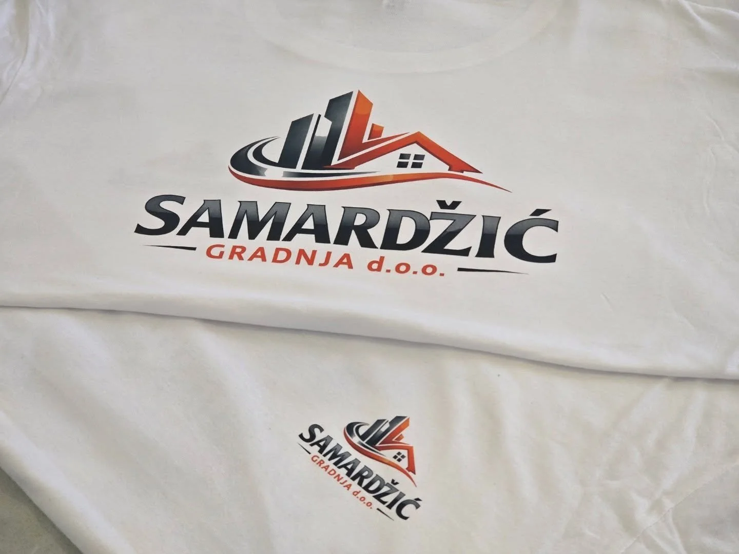 White fabric with a printed logo and text. The logo features stylized buildings, a house, and a curved road in black, gray, and red. Below the logo, the text says "SAMARDŽIĆ" in bold black letters, with "GRADNJA d.o.o." in red underneath.