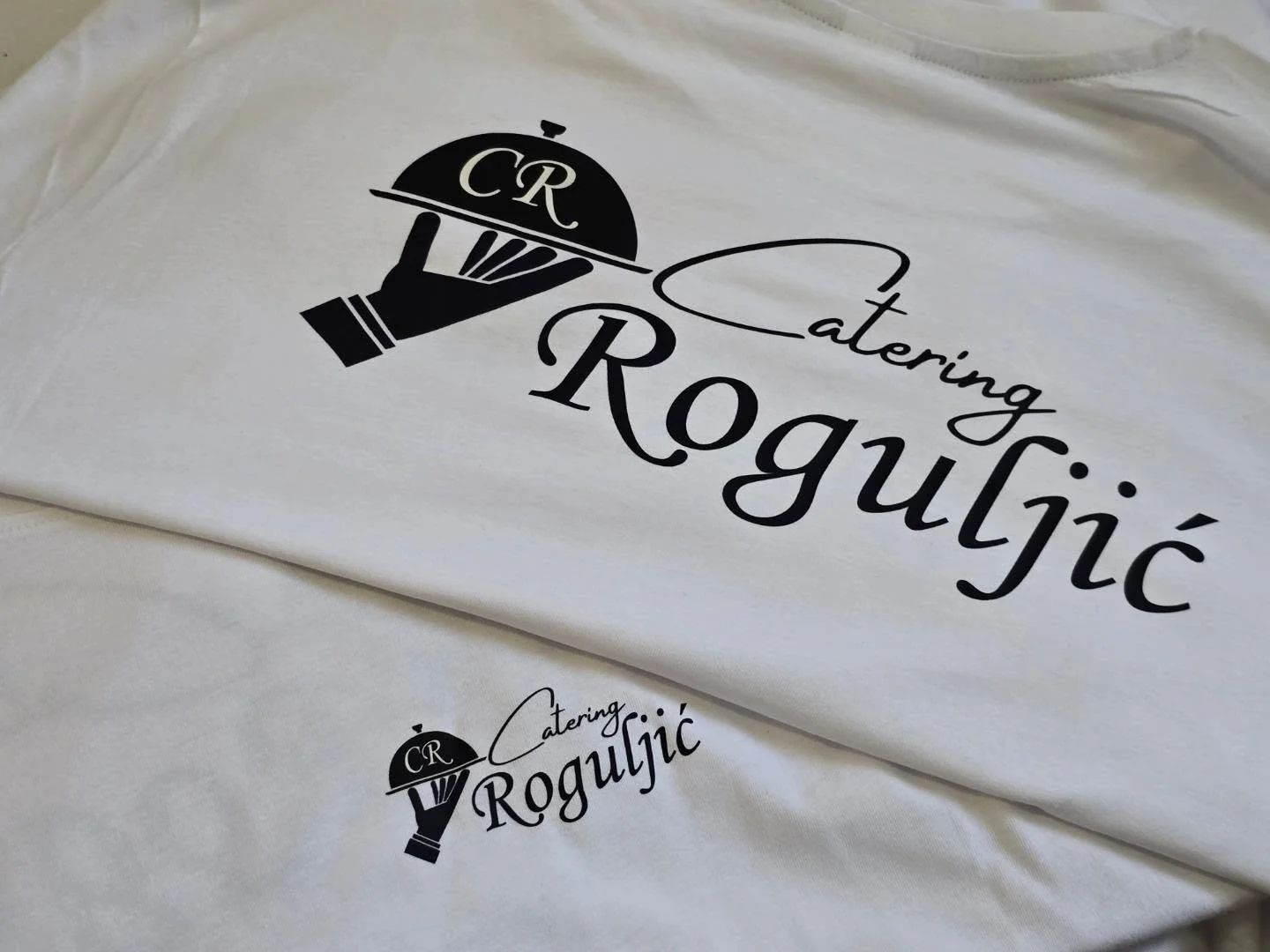 White apron with a black logo and text for 'CR Catering Roguljić' featuring a hand holding a cloche with initials 'CR'.