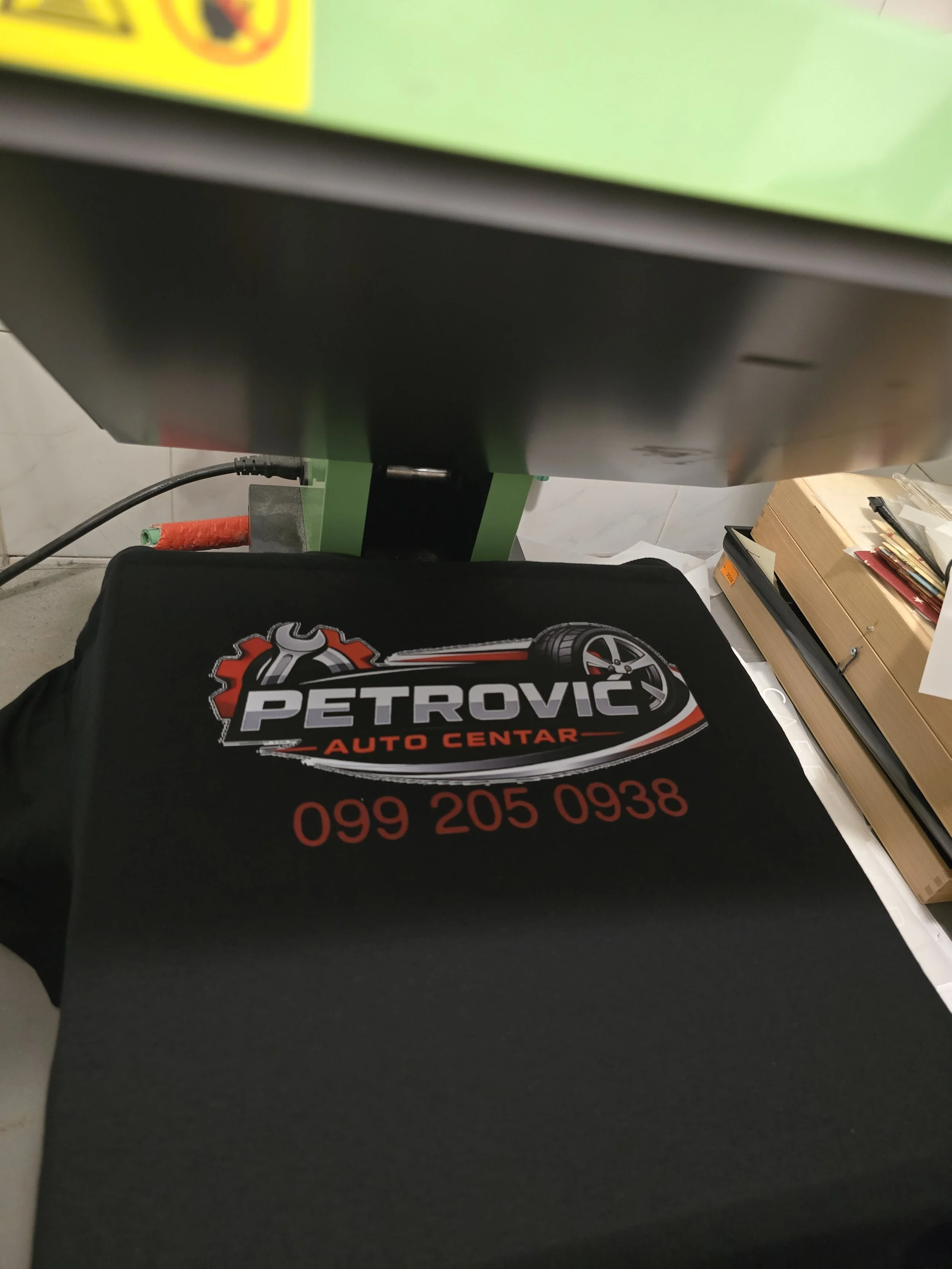 Close-up of a black fabric with a logo and phone number, with a green object and electrical cord partially visible, and boxes with organized papers on the side.