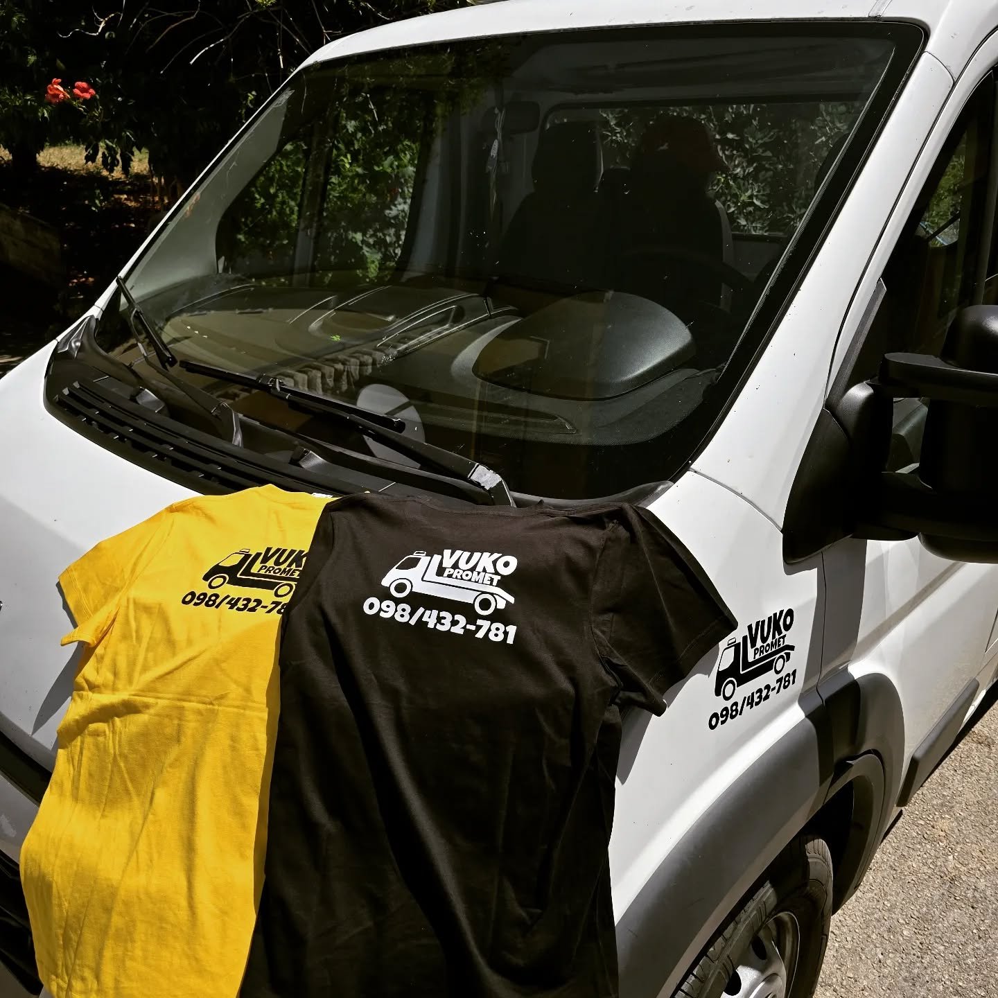 A white utility vehicle with two shirts hanging on its front, one yellow and one black, both bearing a logo and phone number for Vuko Promet, a transportation company. The vehicle is parked outdoors with trees and bushes in the background.