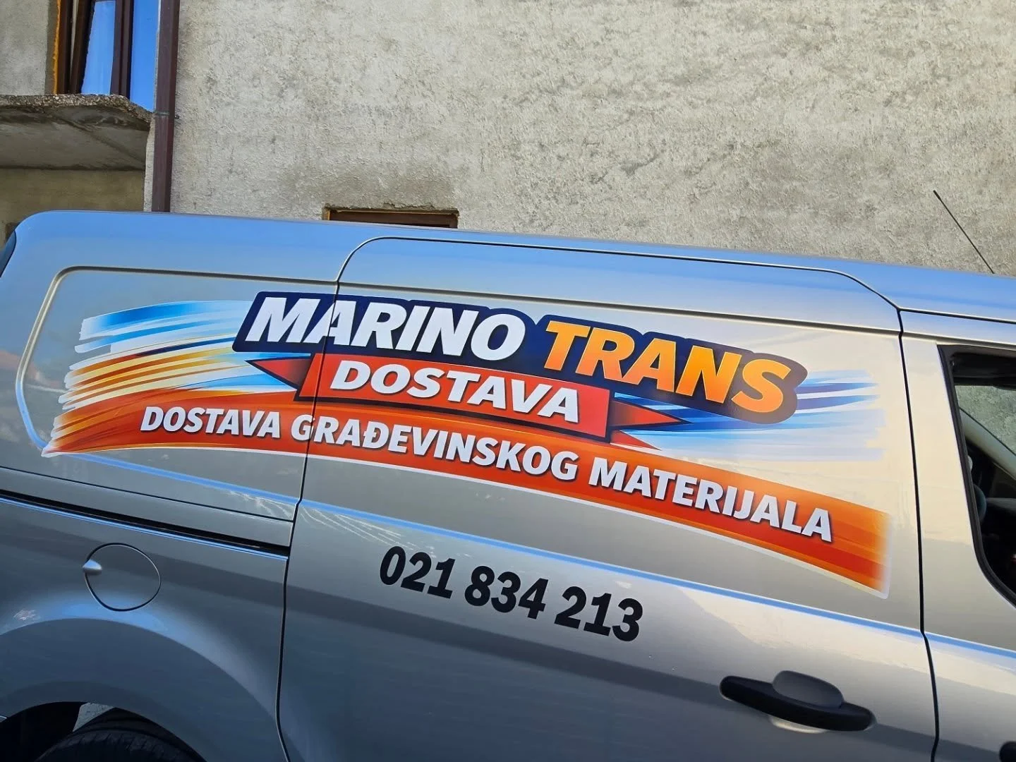 Silver van with colorful advertising decal for Marino Trans, a company delivering building materials. The decal has blue, orange, red, and white text and graphics, including the company's phone number.