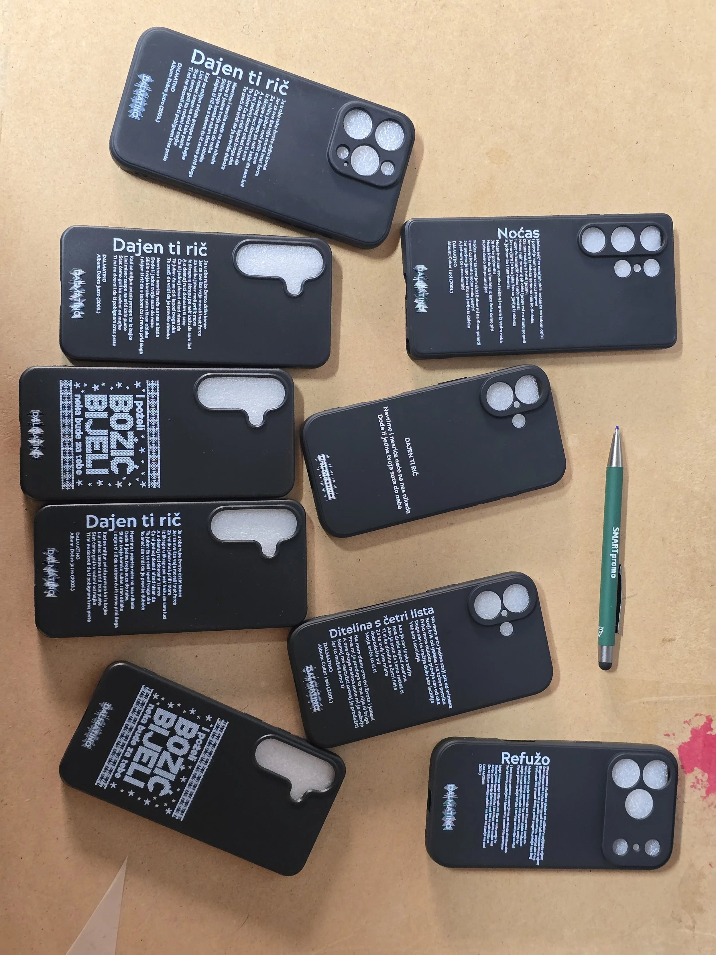 Multiple black smartphone casings with white printed text in Croatian and gray camera cutouts, placed on a beige surface with a green and silver pen nearby.