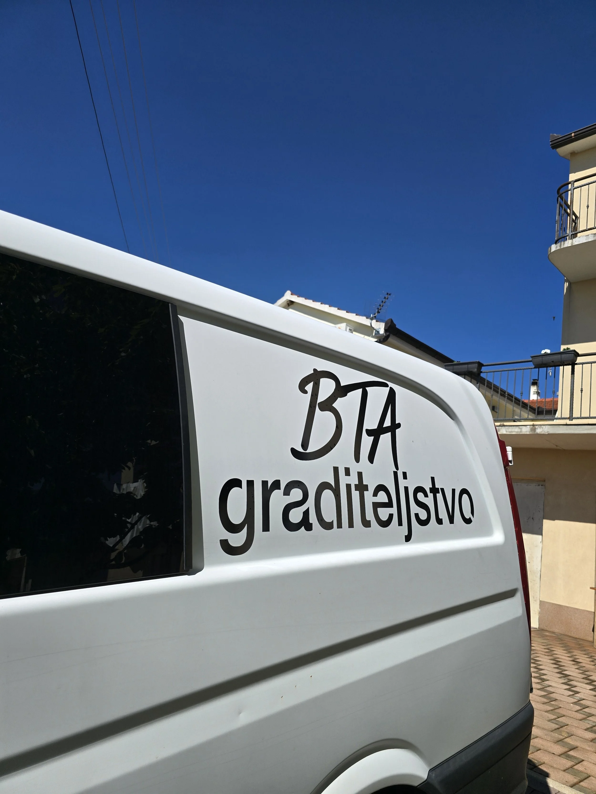 Part of a van with a sign reading 'BIA croissanceistvo' parked on a driveway with a building and balcony in the background, under a clear blue sky.