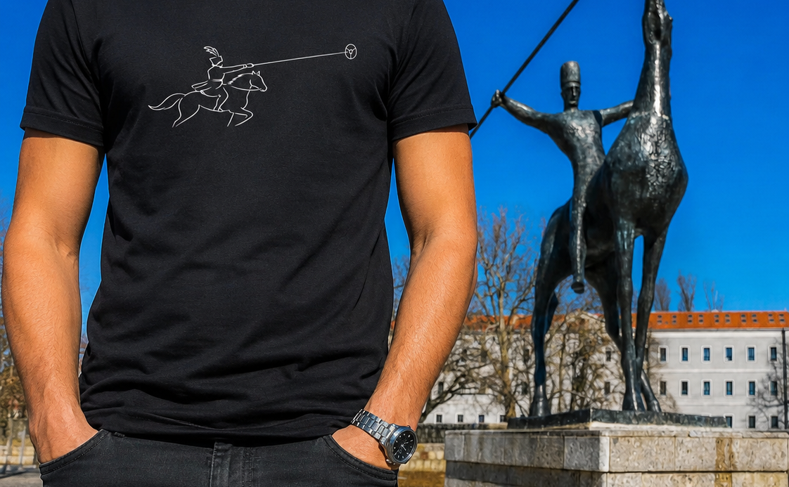 Part of a man wearing a black t-shirt with a graphic of Don Quixote and a windmill, standing in front of a bronze statue of Don Quixote and Sancho Panza riding on a horse, with a background of buildings and leafless trees under a clear blue sky.