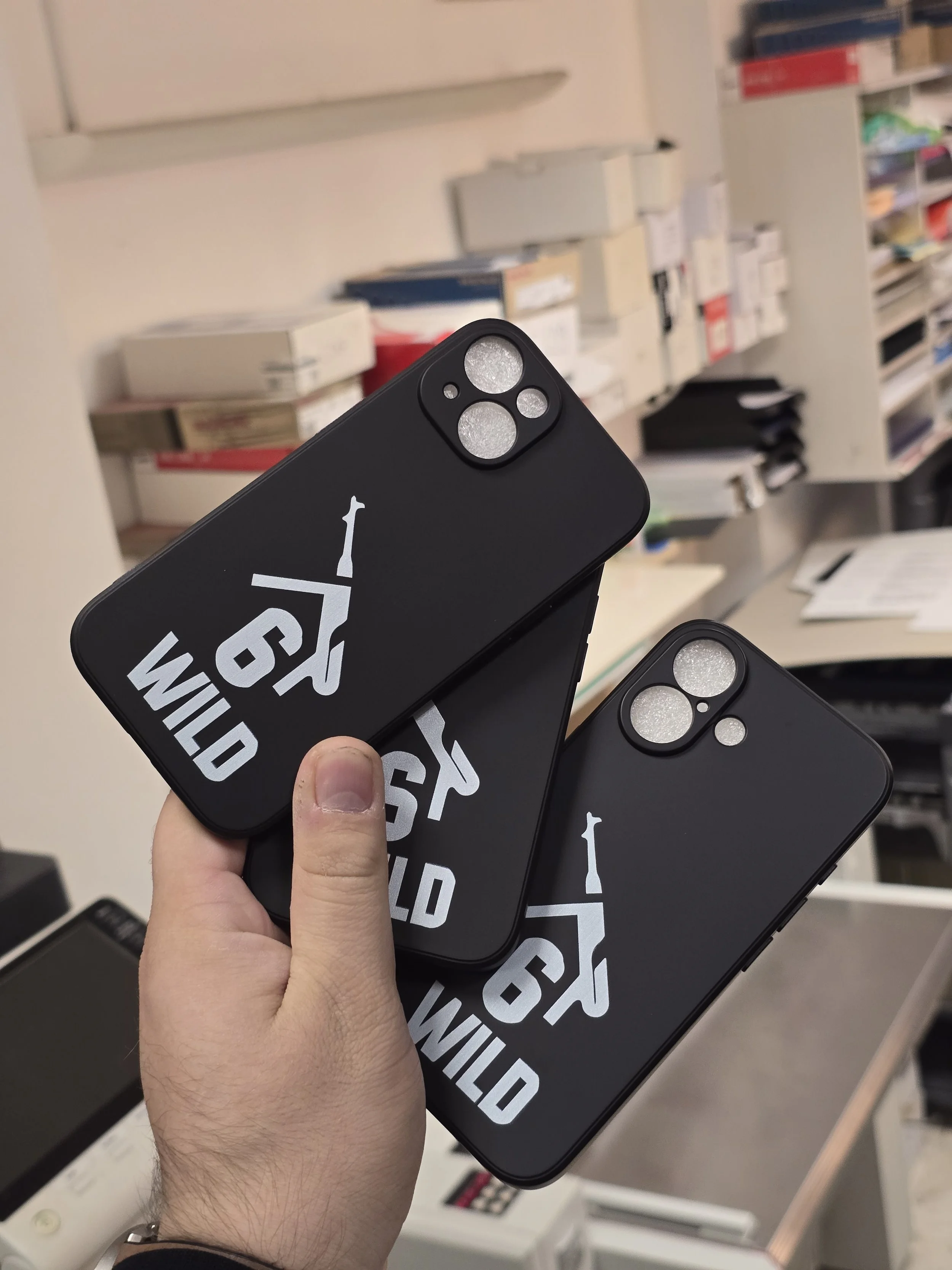 Person holding two smartphones with Wu-Tang Clan logo cases, shelves filled with electronics or accessories in the background.