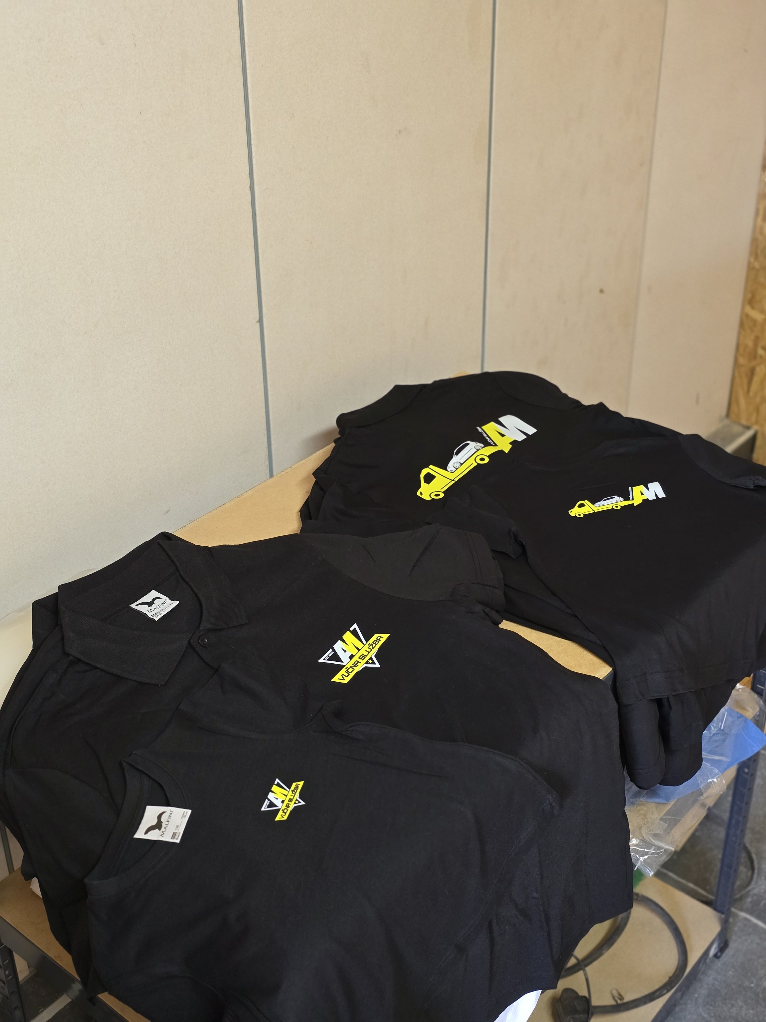 Black t-shirts with a yellow and white logo depicting a car and the text "Vuchna Suzuki" spread on a table in a workshop or store.