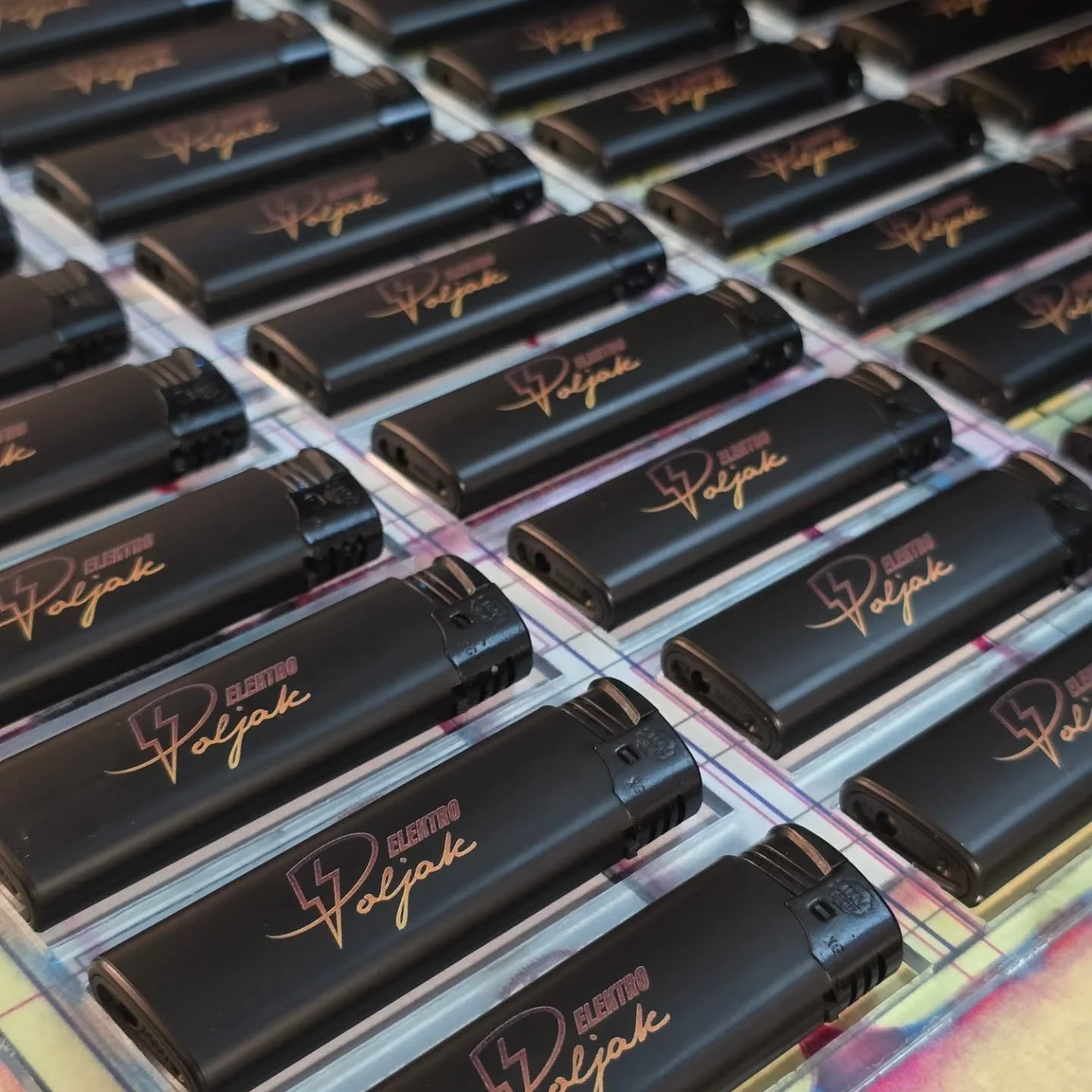 Multiple black lighters with the words 'Electro Vojack' and a lightning bolt design, arranged on a colorful checkered surface.