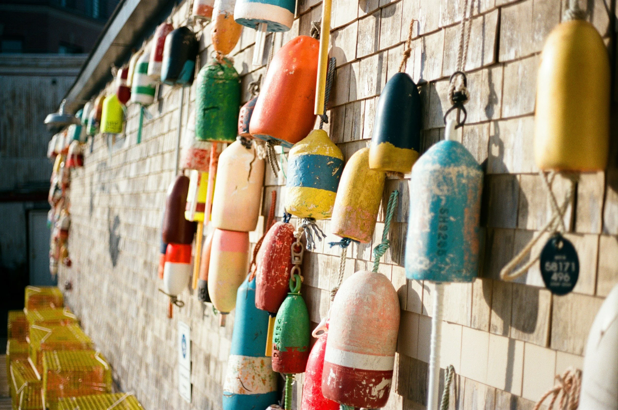 A display of colorful buoys hanging on a wall, with some on the ground, in an outdoor setting with chairs and other objects nearby.