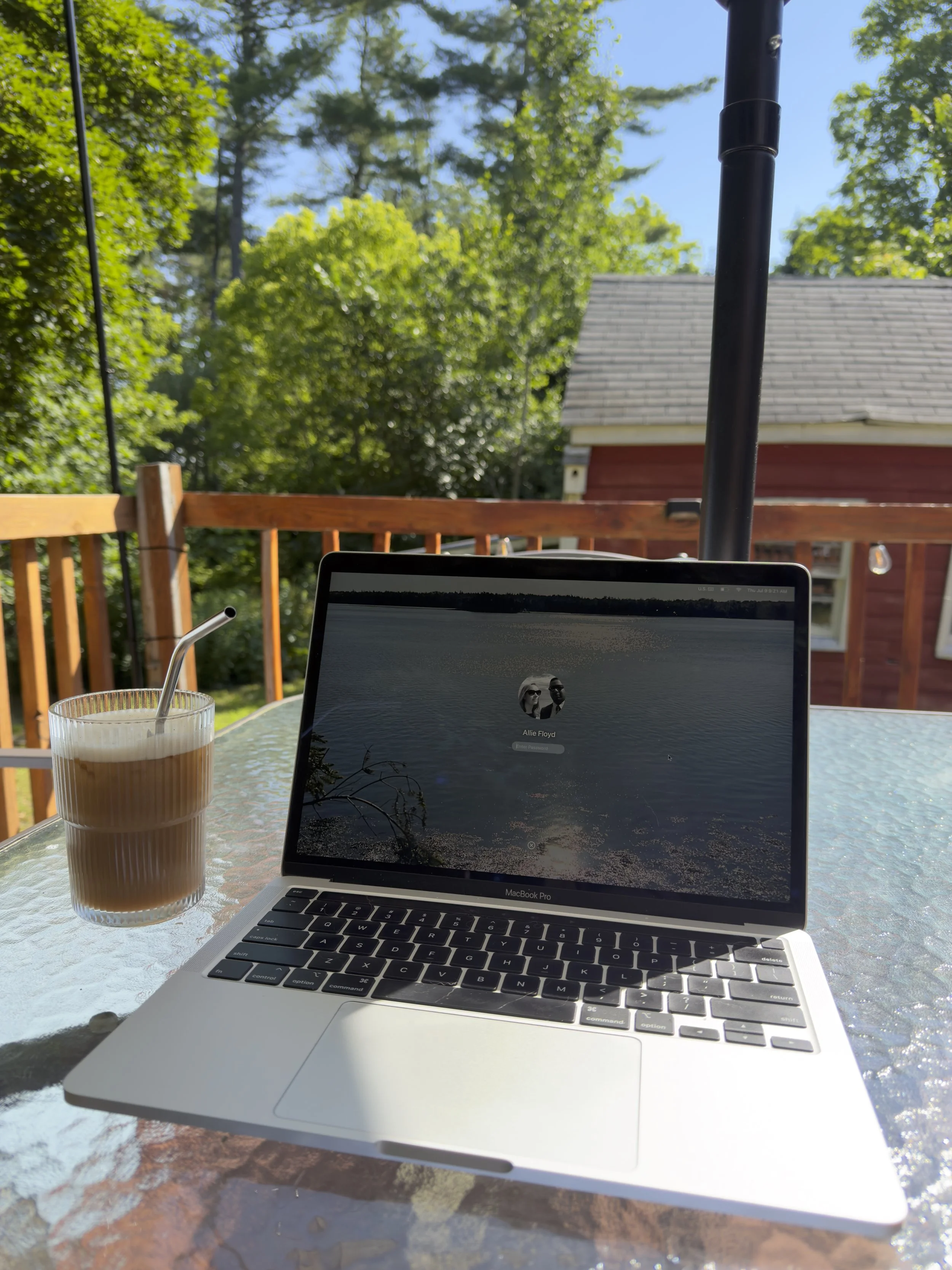 A laptop displaying a login screen with the name Allie Floyd is on a glass-topped table outdoors, accompanied by a tall glass of iced coffee with a straw, on a patio with a wooden railing, surrounded by green trees, with a house with gray roofing in the background and a bright blue sky.