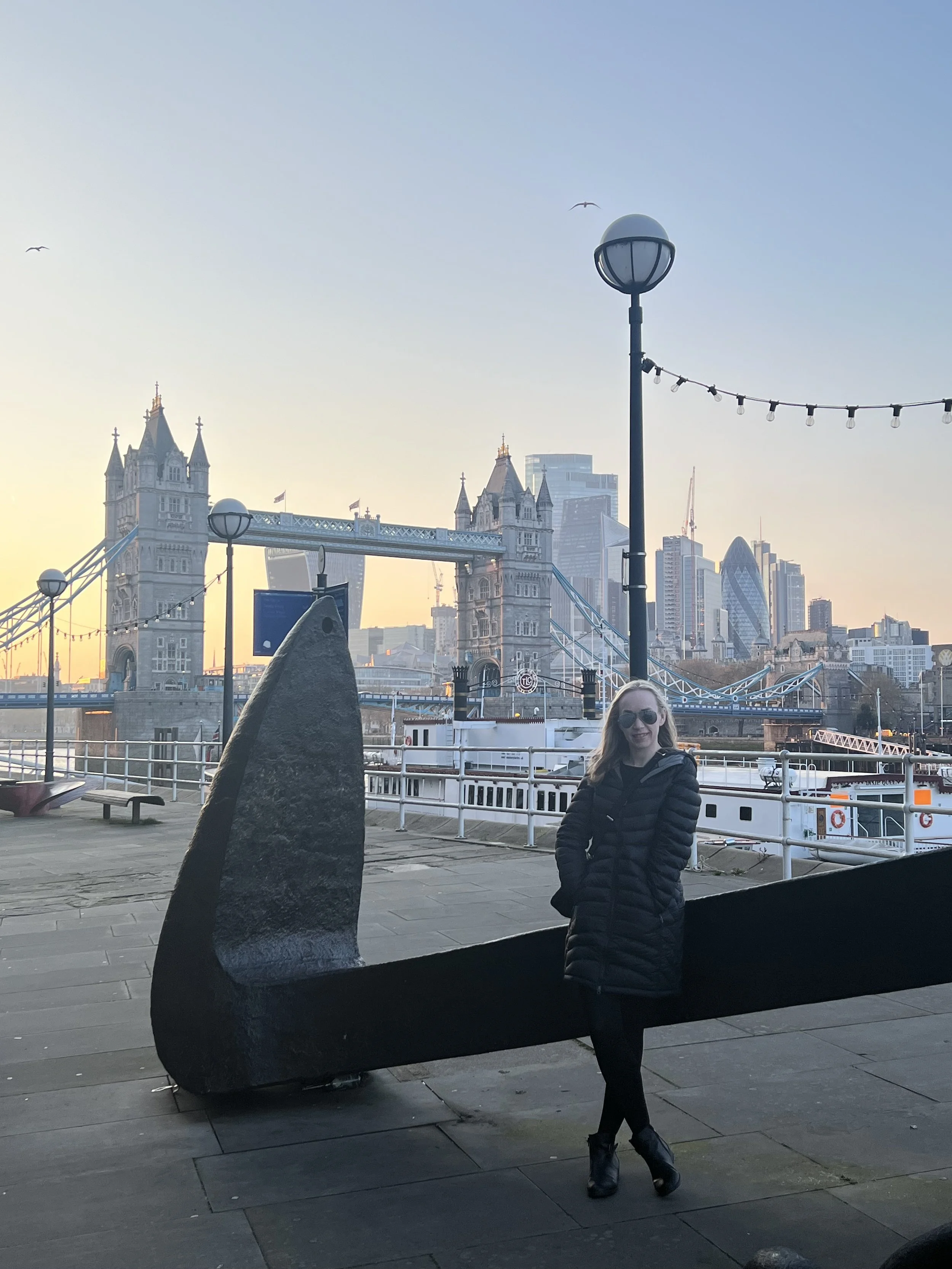 A woman with blonde hair, wearing sunglasses and a black puffer jacket, standing outdoors near a large black sculpture of the letter L. In the background, the Tower Bridge in London is visible with its twin towers and suspension cables, along with modern skyscrapers. The sky is clear with some birds flying in the distance.