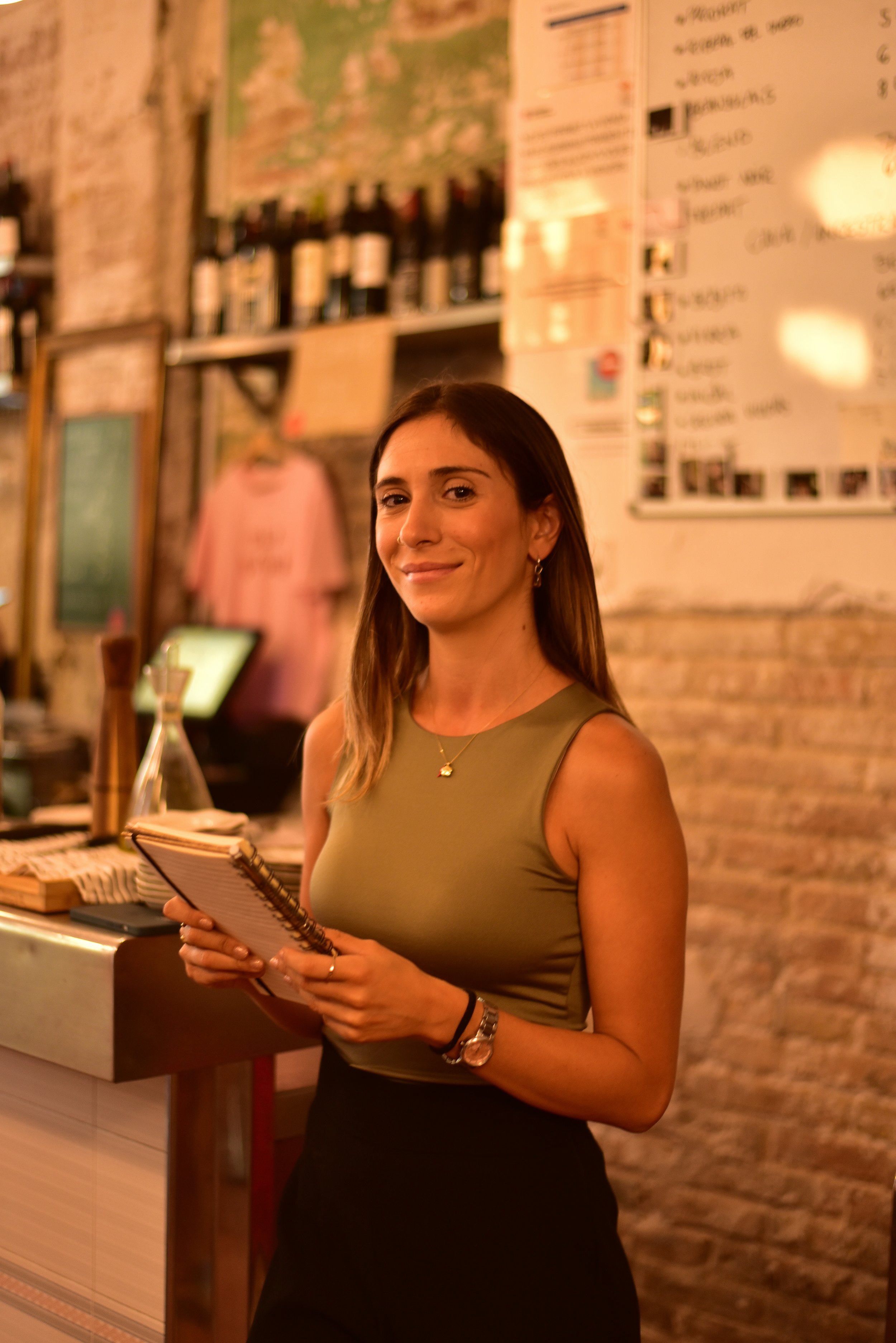 Mujer sonriendo, sosteniendo un cuaderno, en un restaurante con paredes de ladrillo y un menú visible en el fondo.
