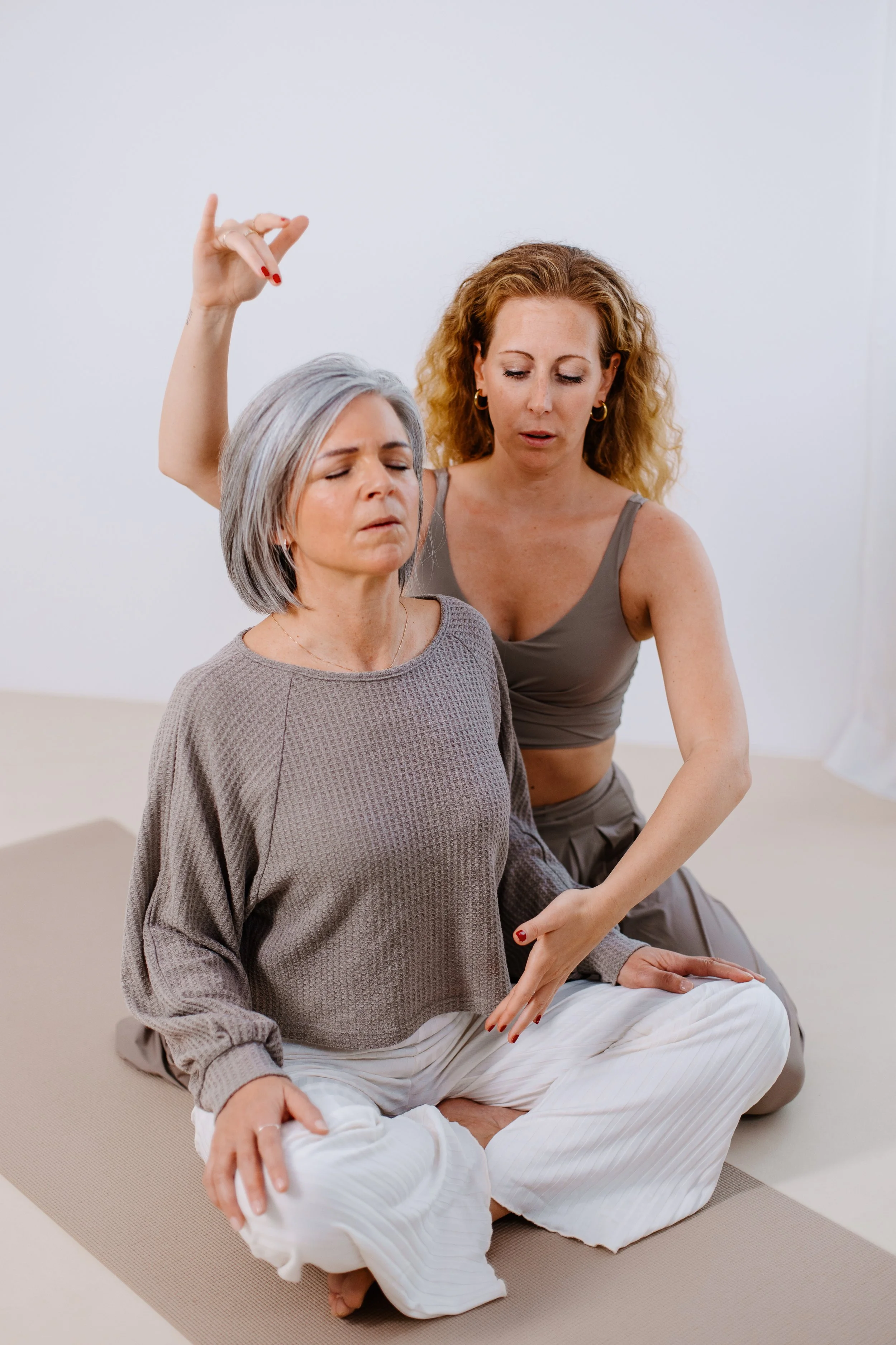 Drei Frauen üben Yoga auf einer Matte in einem hellen Raum. Die ältere Frau sitzt im Schneidersitz mit geschlossenen Augen, die jüngere Frau macht eine meditative Geste hinter ihr.