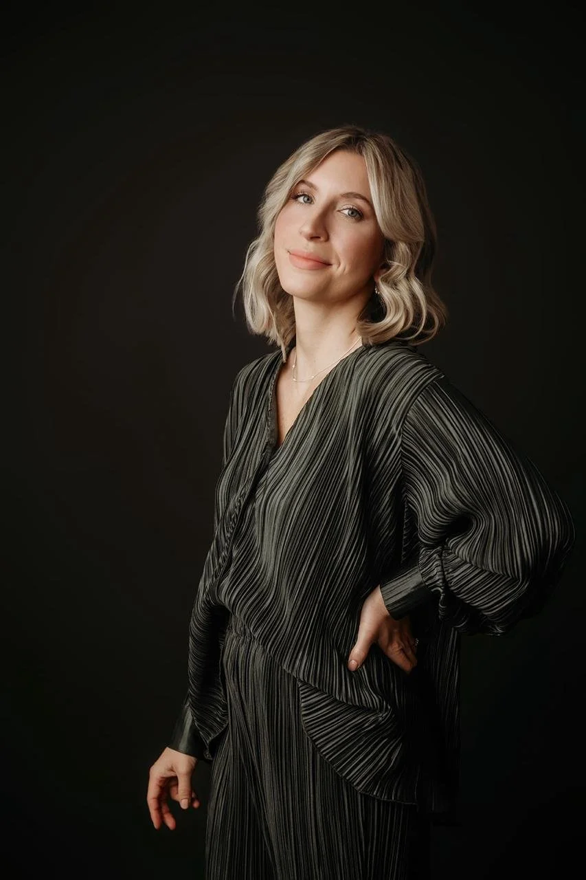 Ein Portrait einer lächelnden Frau mit blonden, gewellten Haaren, die eine schwarze, gestreifte Bluse trägt, vor dunklem Hintergrund.