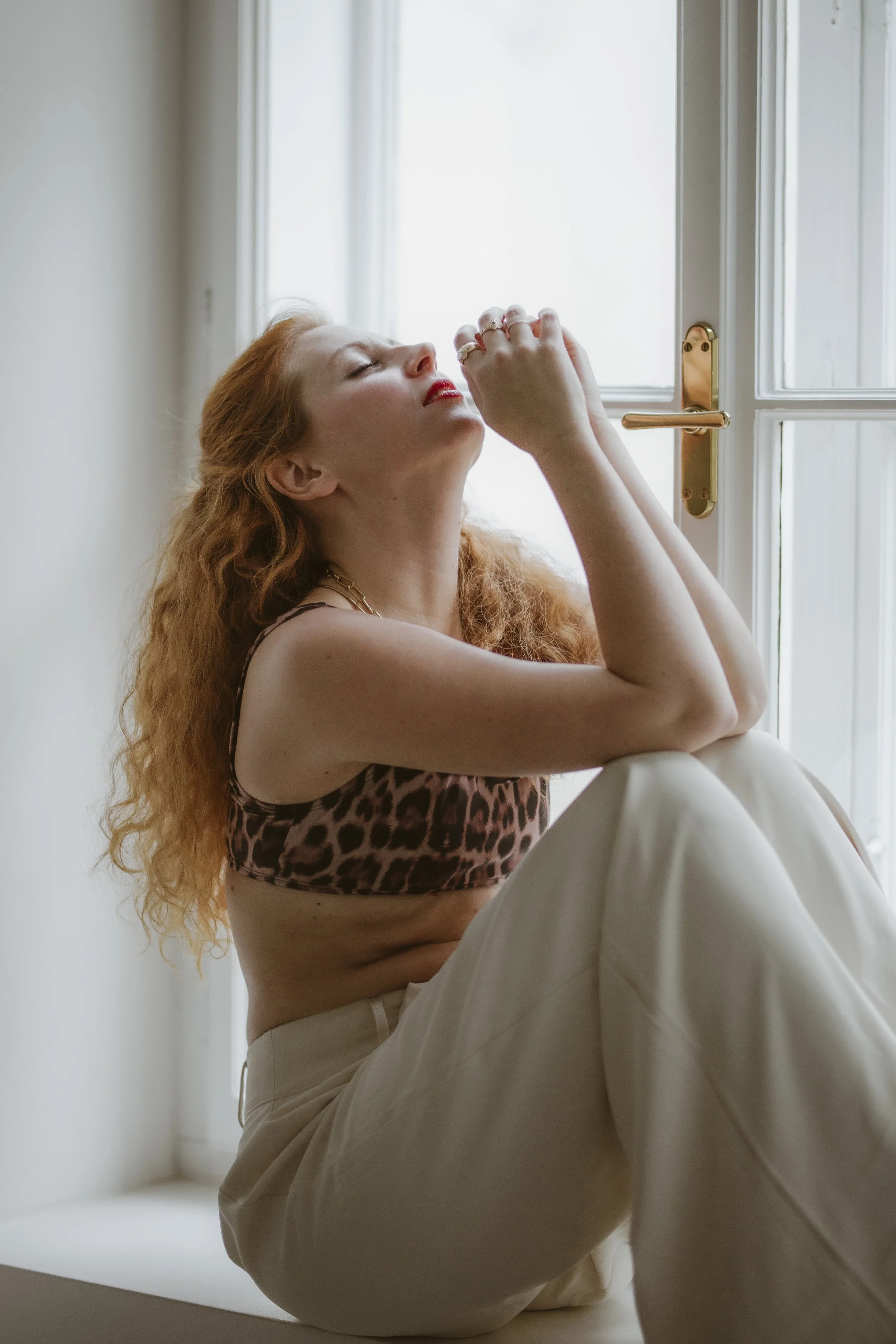 Eine Frau mit roten, lockigen Haaren sitzt an einem Fenster, die Augen geschlossen, trägt ein leopardmusterndes Oberteil und eine weiße Hose.