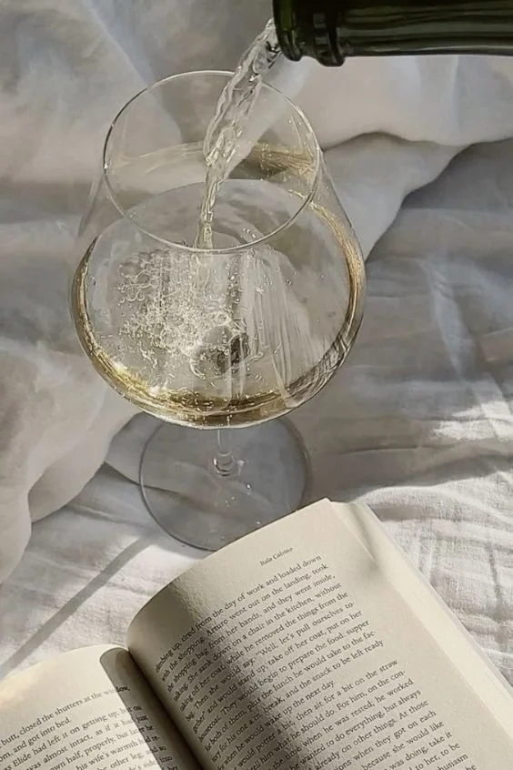 Ein Weinglas, aus dem Weißwein eingeschenkt wird, liegt auf einem weißen Tuch neben einem aufgeschlagenen Buch.