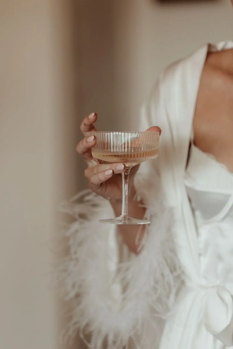 Frau in eleganter weißer Robe hält ein Kristall-Getränkeglas mit Champagner