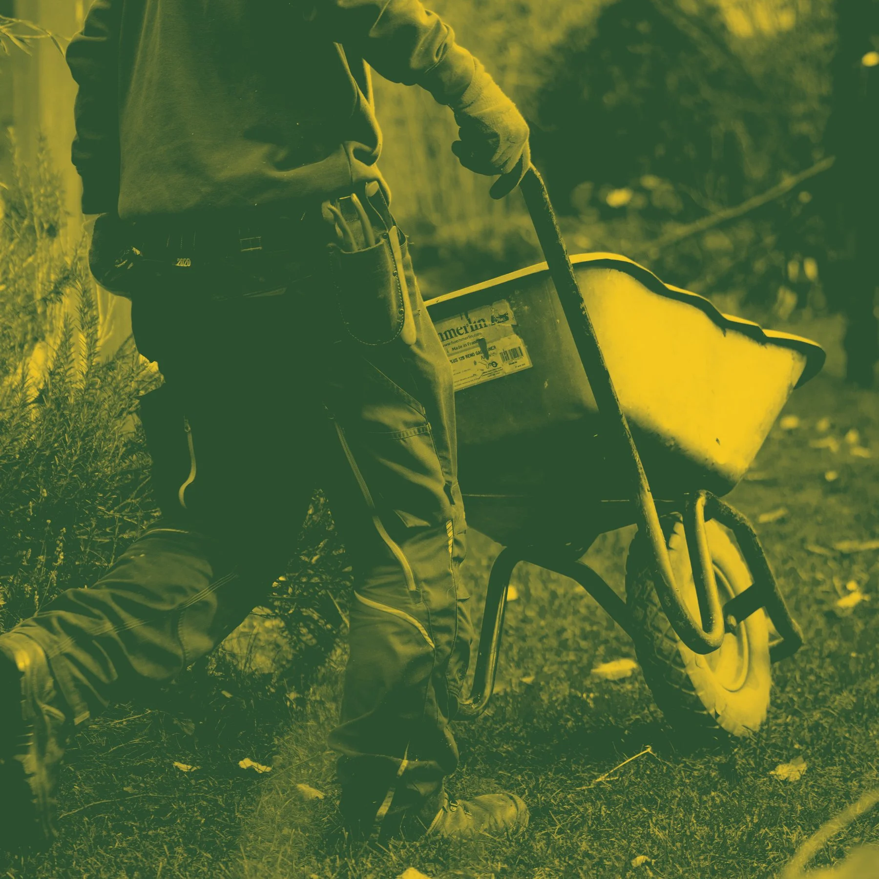 Close-up of a person wearing a hoodie and long pants, holding a yellow wheelbarrow outside on grass with trees in the background.