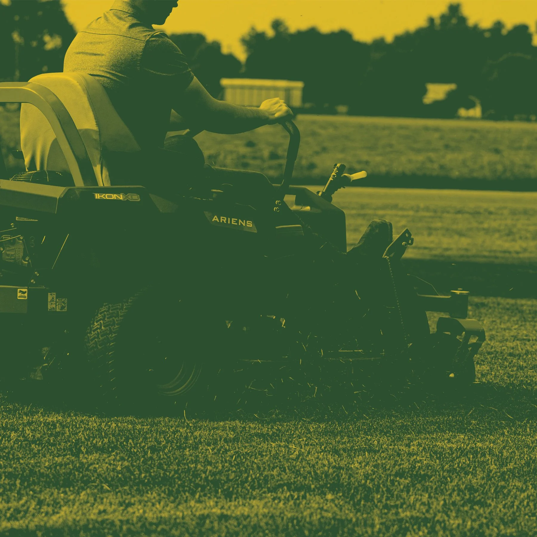 A person riding a green all-terrain vehicle (ATV) on a grassy field with trees and buildings in the background during sunset.