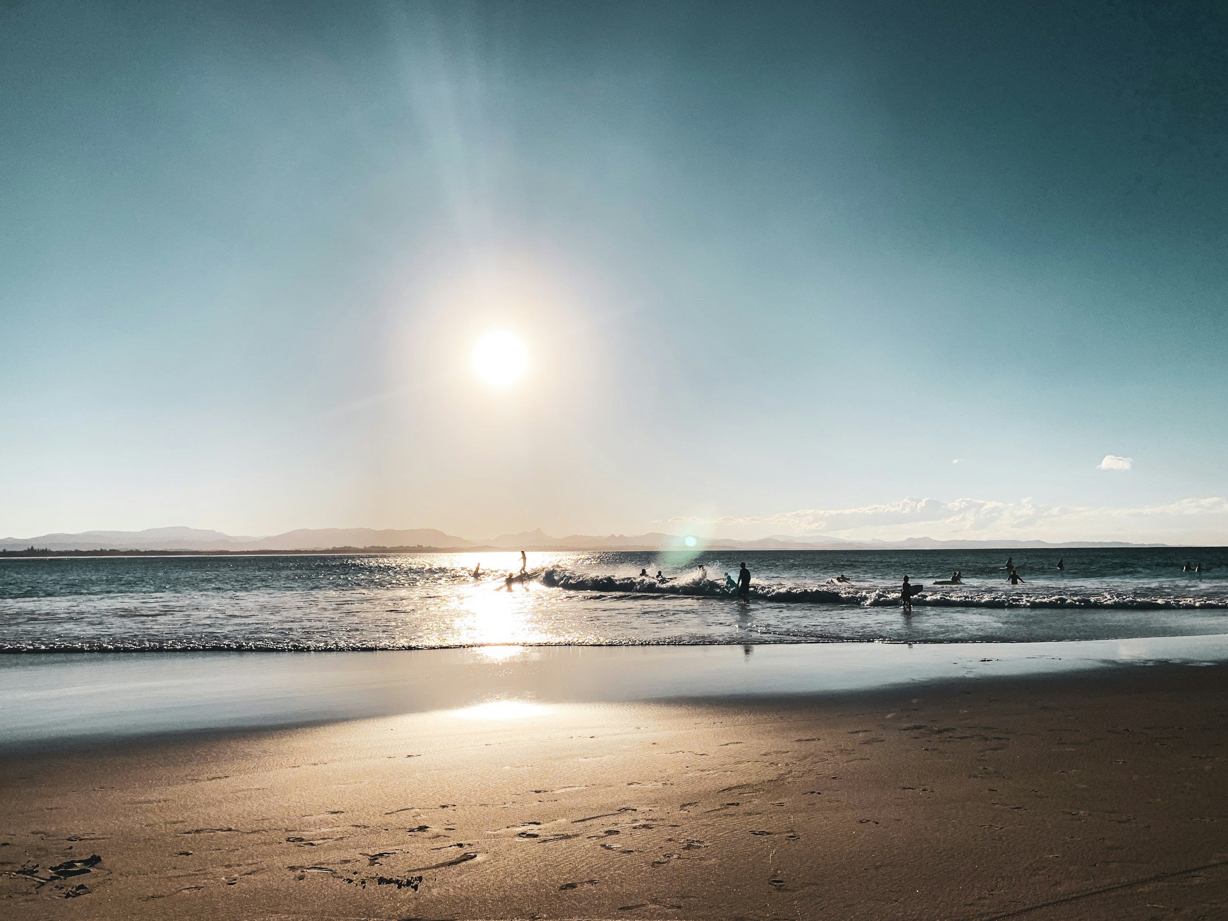 Plage au coucher de soleil avec des personnes nageant et jouant dans l'eau, horizon montagneux visible au loin, ciel clair et soleil brillant.