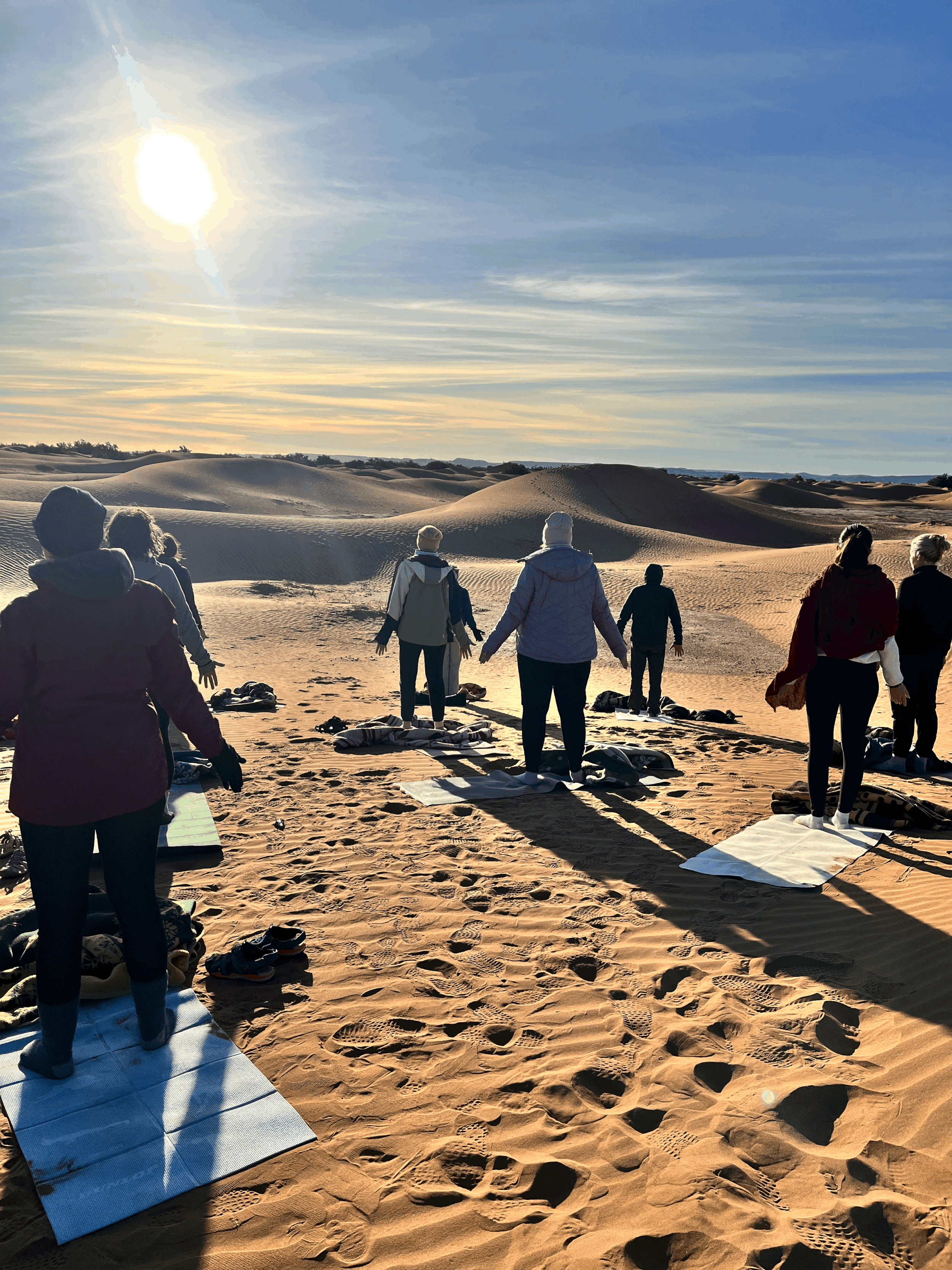 Groupe de personnes pratiquant la méditation ou le yoga sur un tapis dans un désert au lever du soleil.