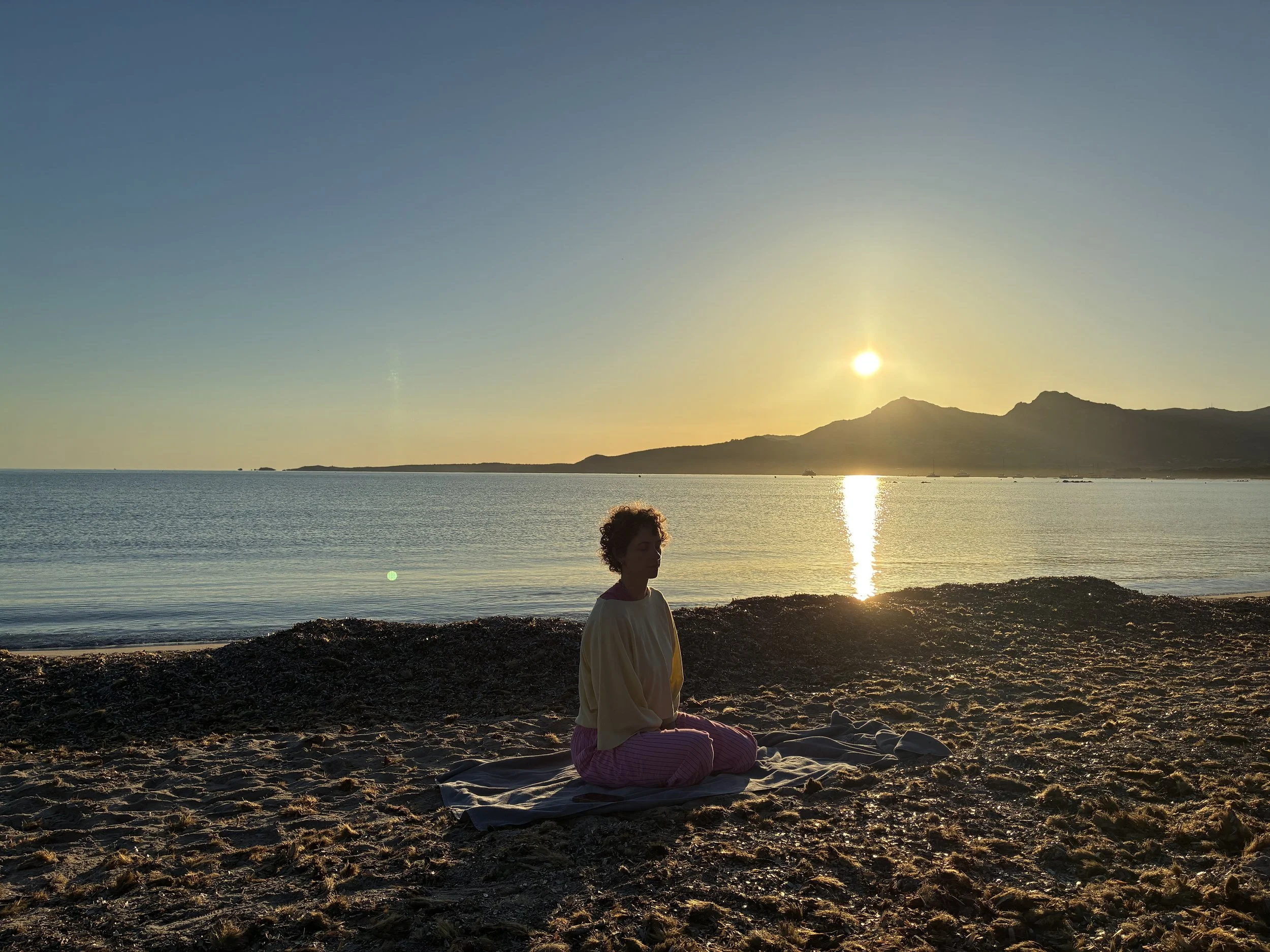 Une personne assise au bord de la mer, contemplant un coucher de soleil derrière des montagnes, avec une lumière douce et une ambiance paisible.
