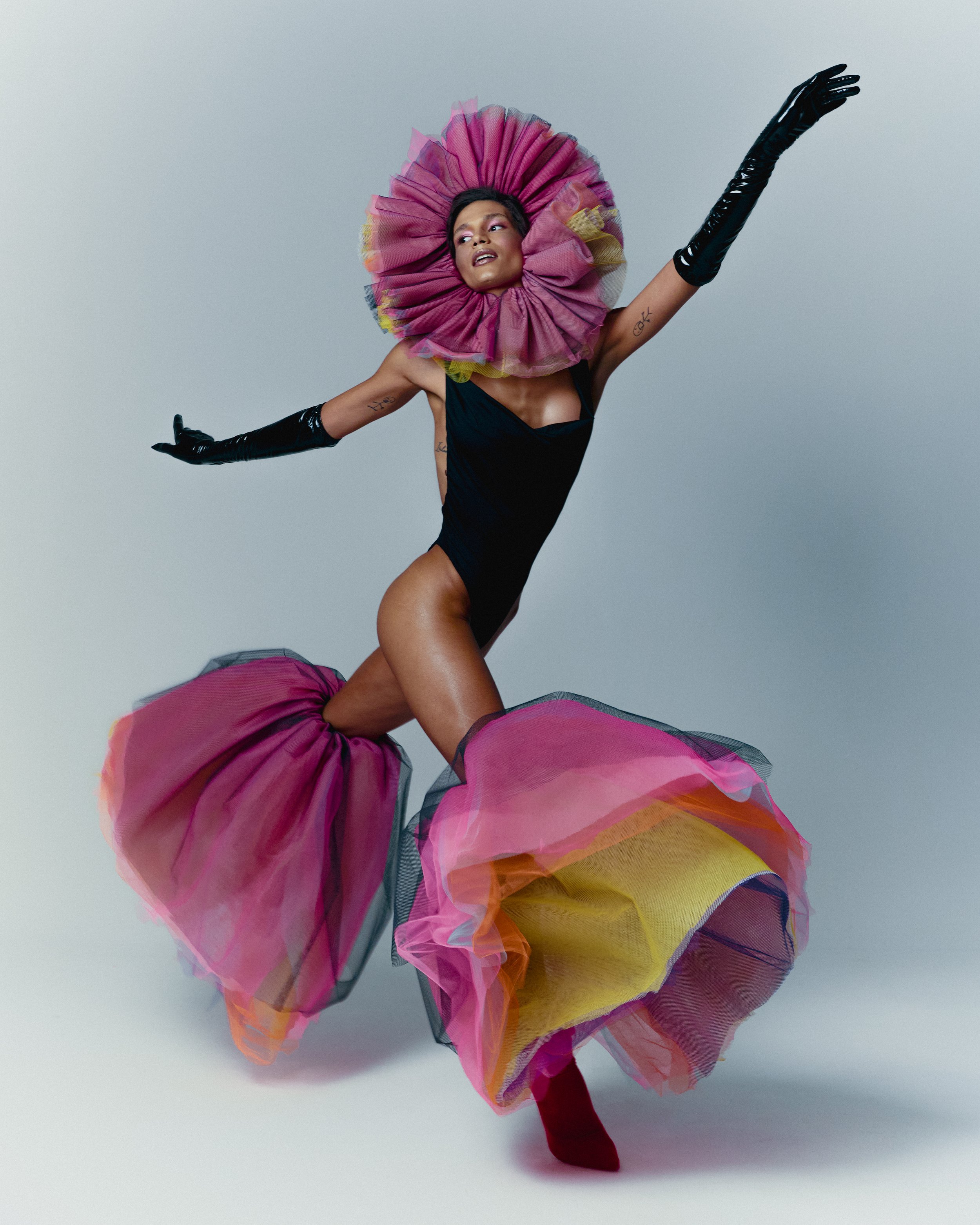 Figura de uma pessoa com pele escura, vestida com traje de balé preto, usando uma saia de tule colorido em tons de rosa, amarelo e laranja, com uma cabeça decorada com uma grande gola de tule rosa com detalhes em amarelo, fazendo pose de dança contra fundo cinza