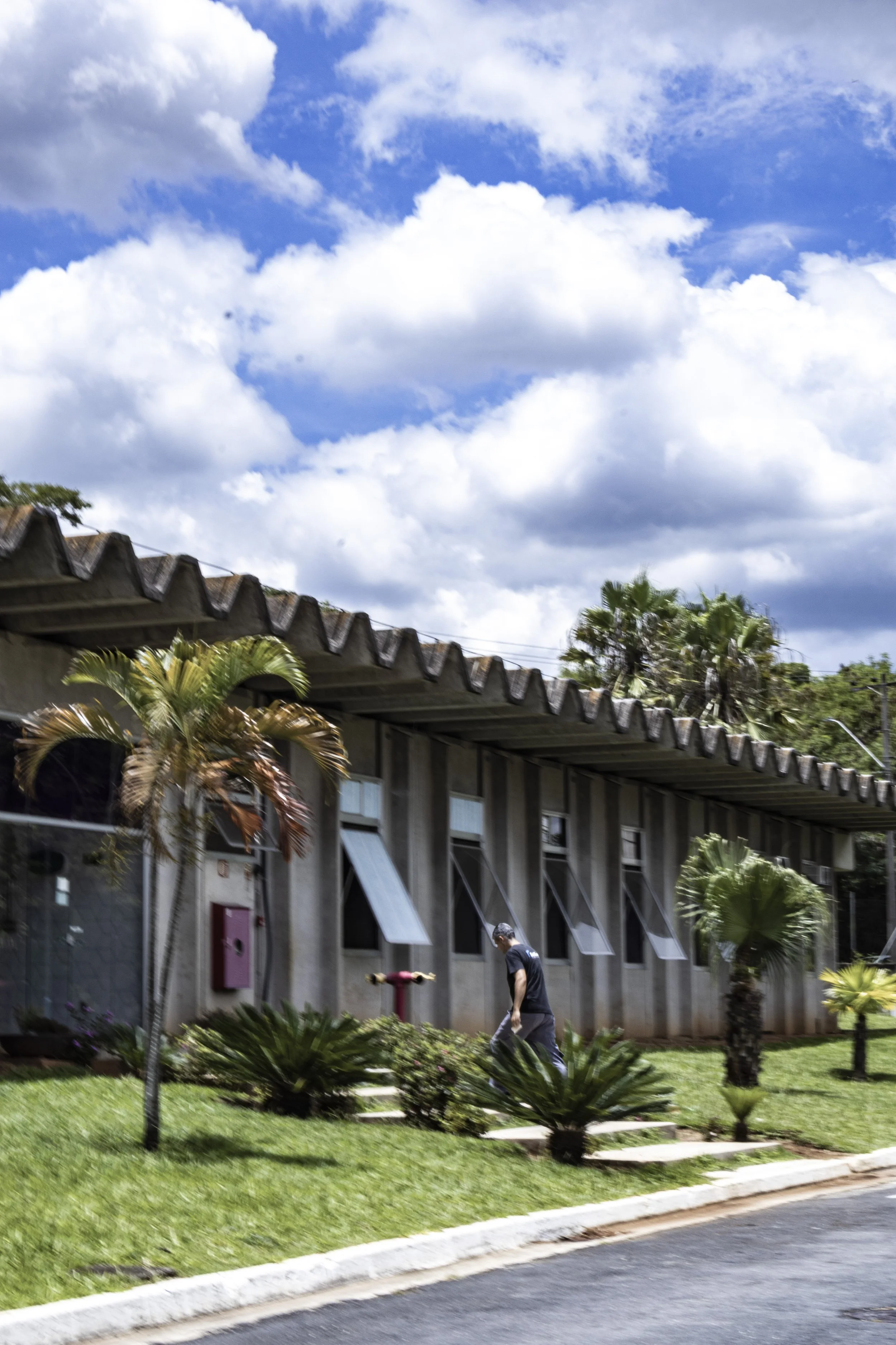 Edifício de vários andares com janelas retangulares, cercado de árvores e plantas ornamentais, sob céu ensolarado com nuvens brancas.
