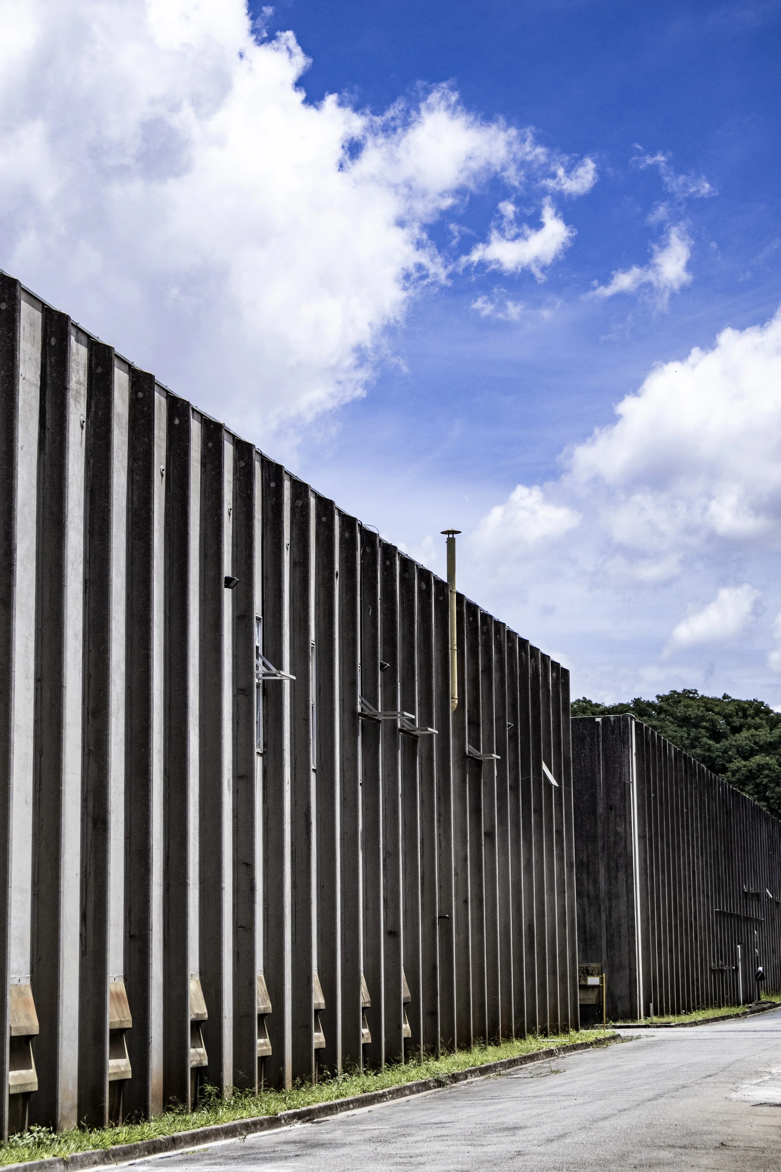 Muro de concreto com painéis verticais e uma grade perto da calçada, com céu azul com nuvens brancas ao fundo.