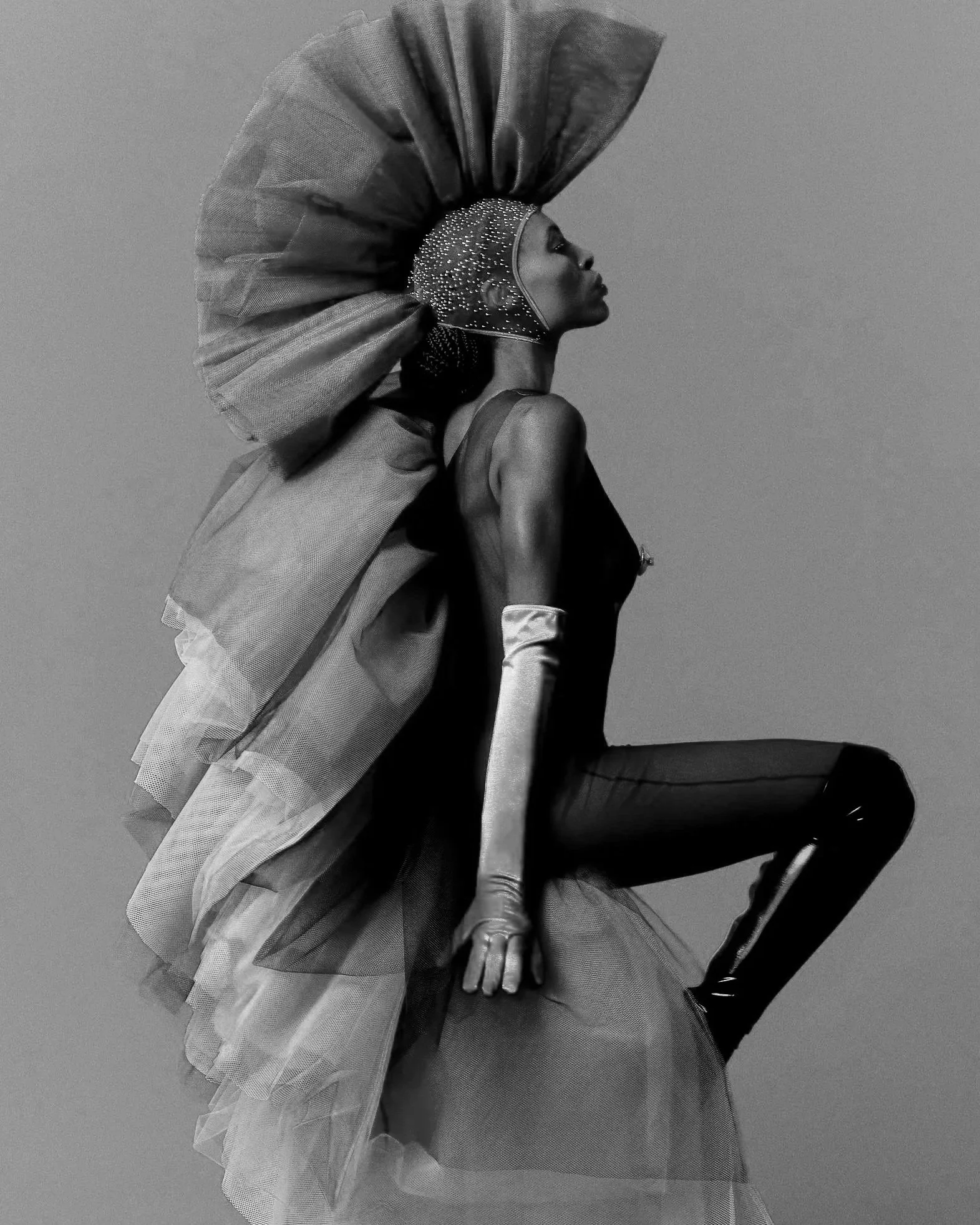 Mulher usando fantasia futurista com cabeça coberta por pequenos pontos luminosos, vestindo uma jaqueta e uma saia de tule, sentada em uma cadeira.