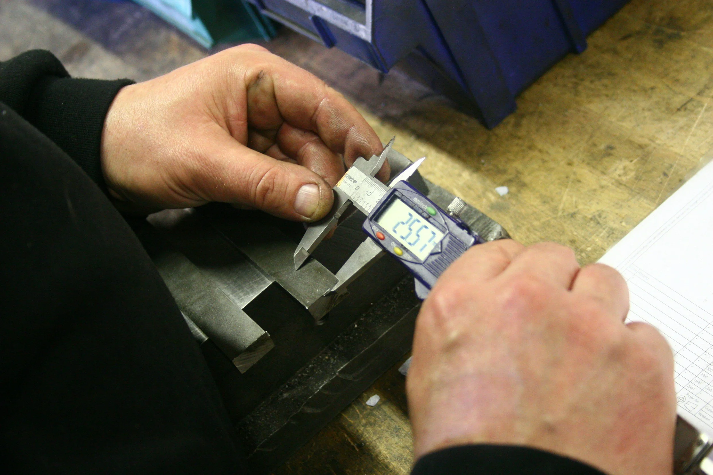 A person uses a digital caliper to measure the width of a metal piece on a workbench, with a background of workshop items and paper.