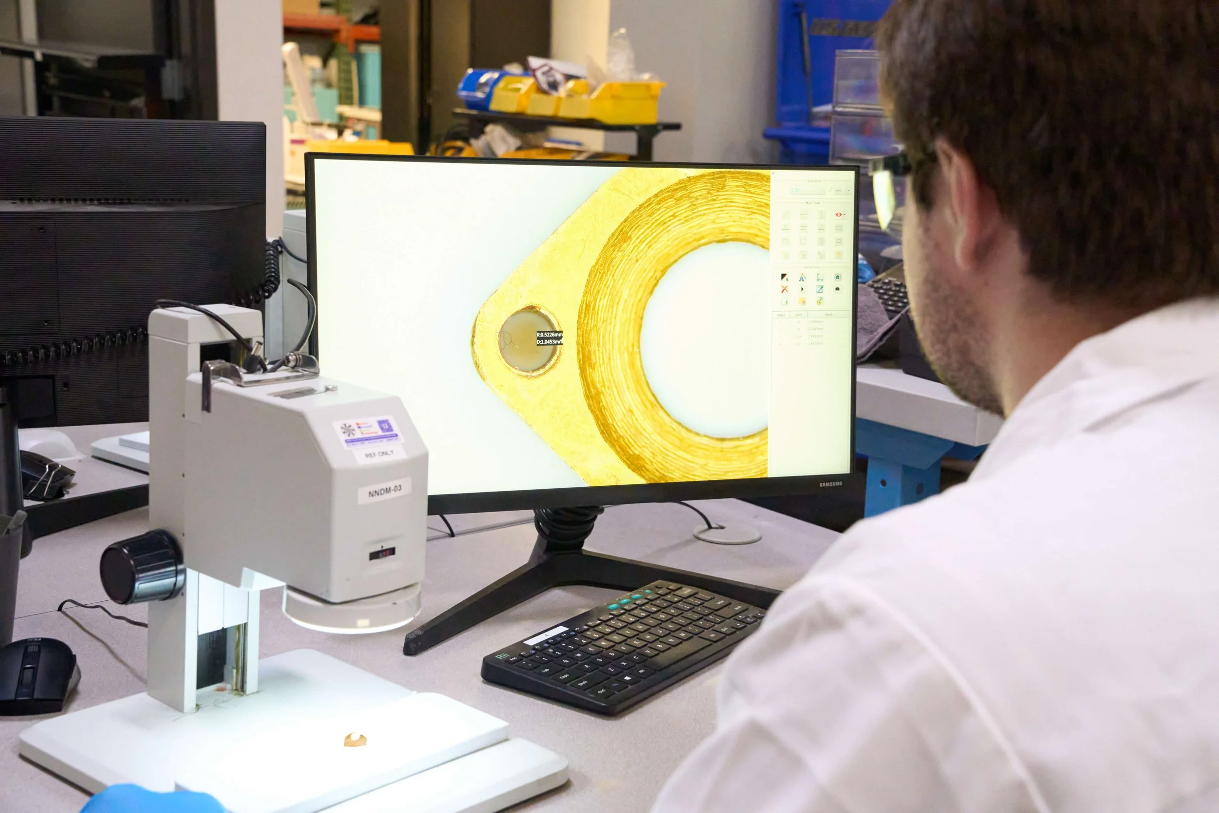 A person in a laboratory or research setting is using a machine connected to a computer, viewing an image of a cross-section of a material on the screen. The setup includes industrial equipment and shelves with boxes in the background.