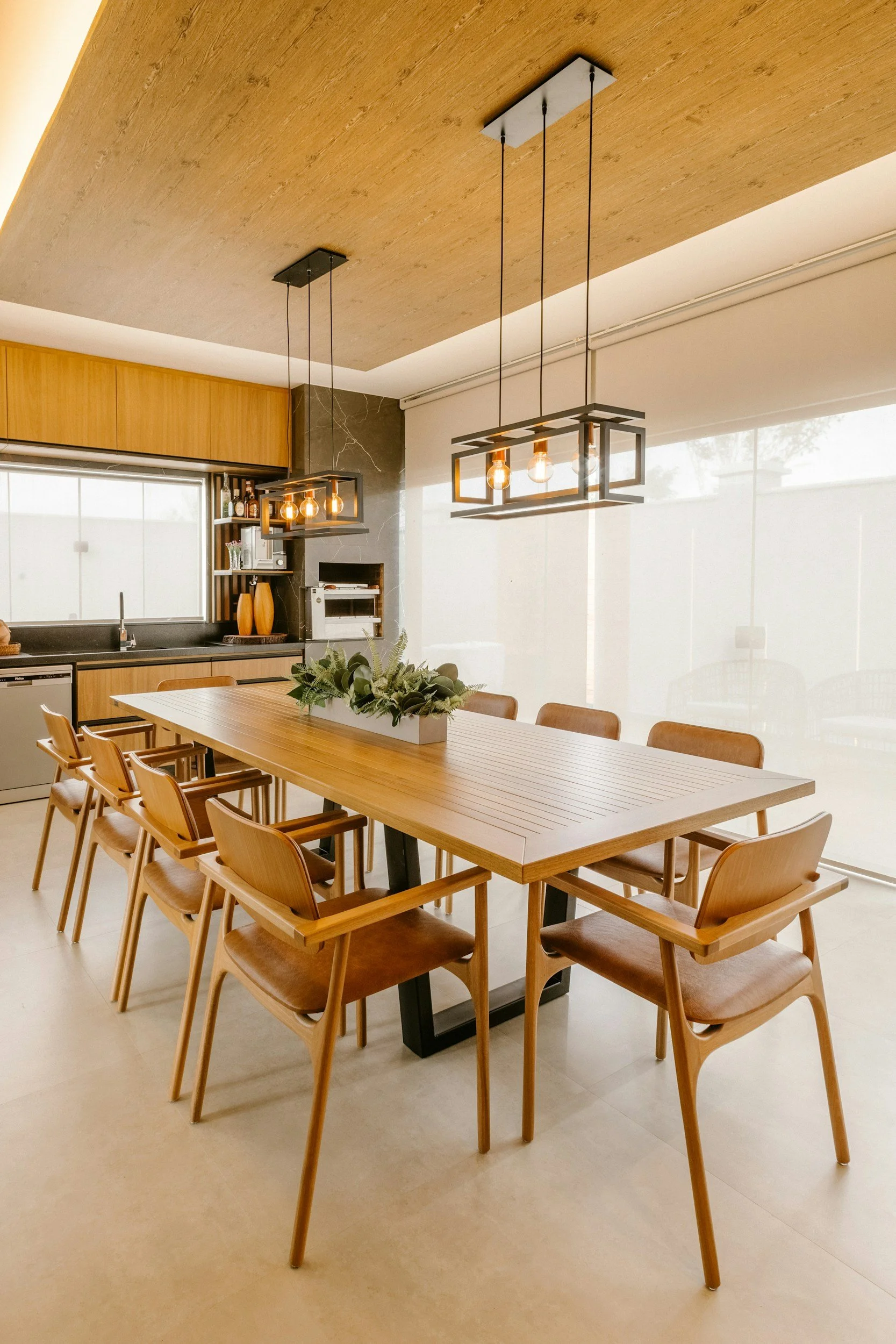 Modern dining room with a long wooden table surrounded by matching wooden chairs, a ceiling with wooden panels, and three contemporary pendant lights hanging above.