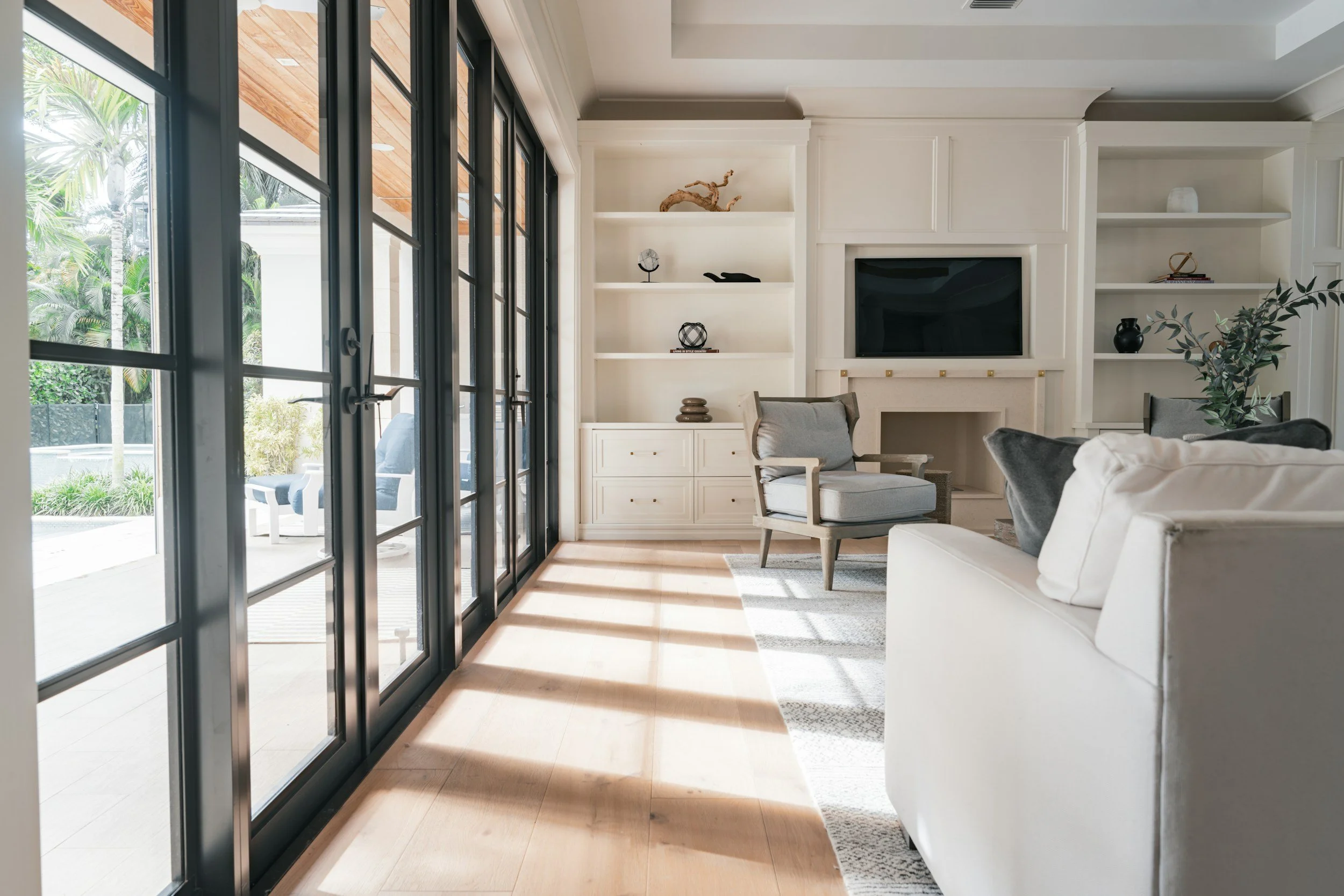 Living room with large black-framed glass doors opening to a patio, white built-in shelves with decor, a television, light-colored armchair, sofa, and a large plant.