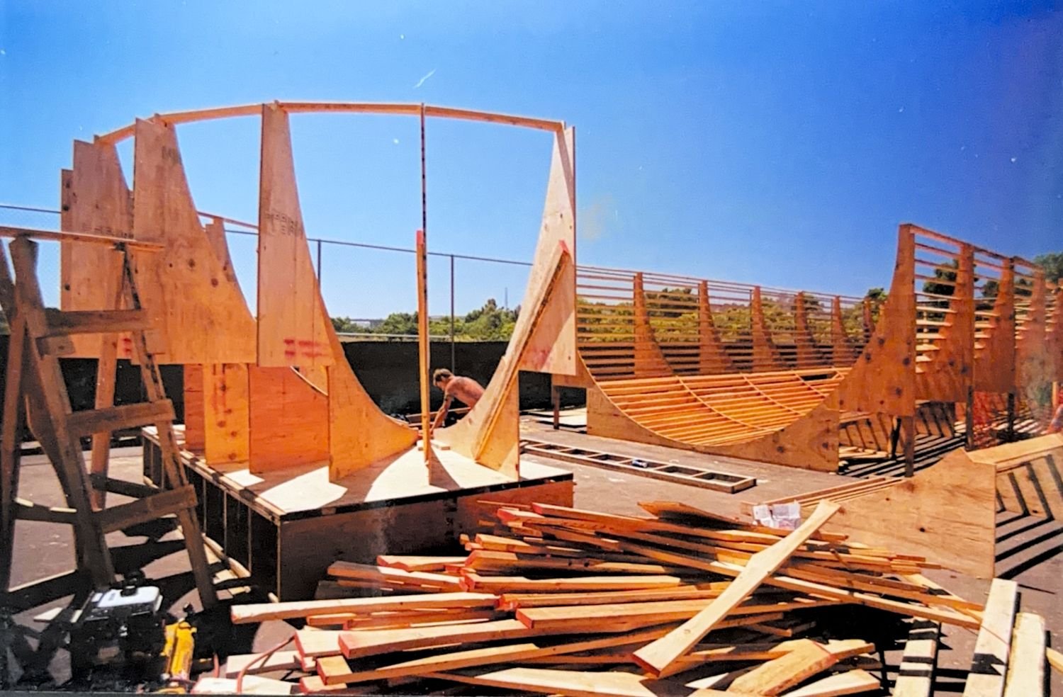 Wooden skateboarding ramp under construction with scattered wood pieces and tools, a worker assembling it outdoors on a bright sunny day.
