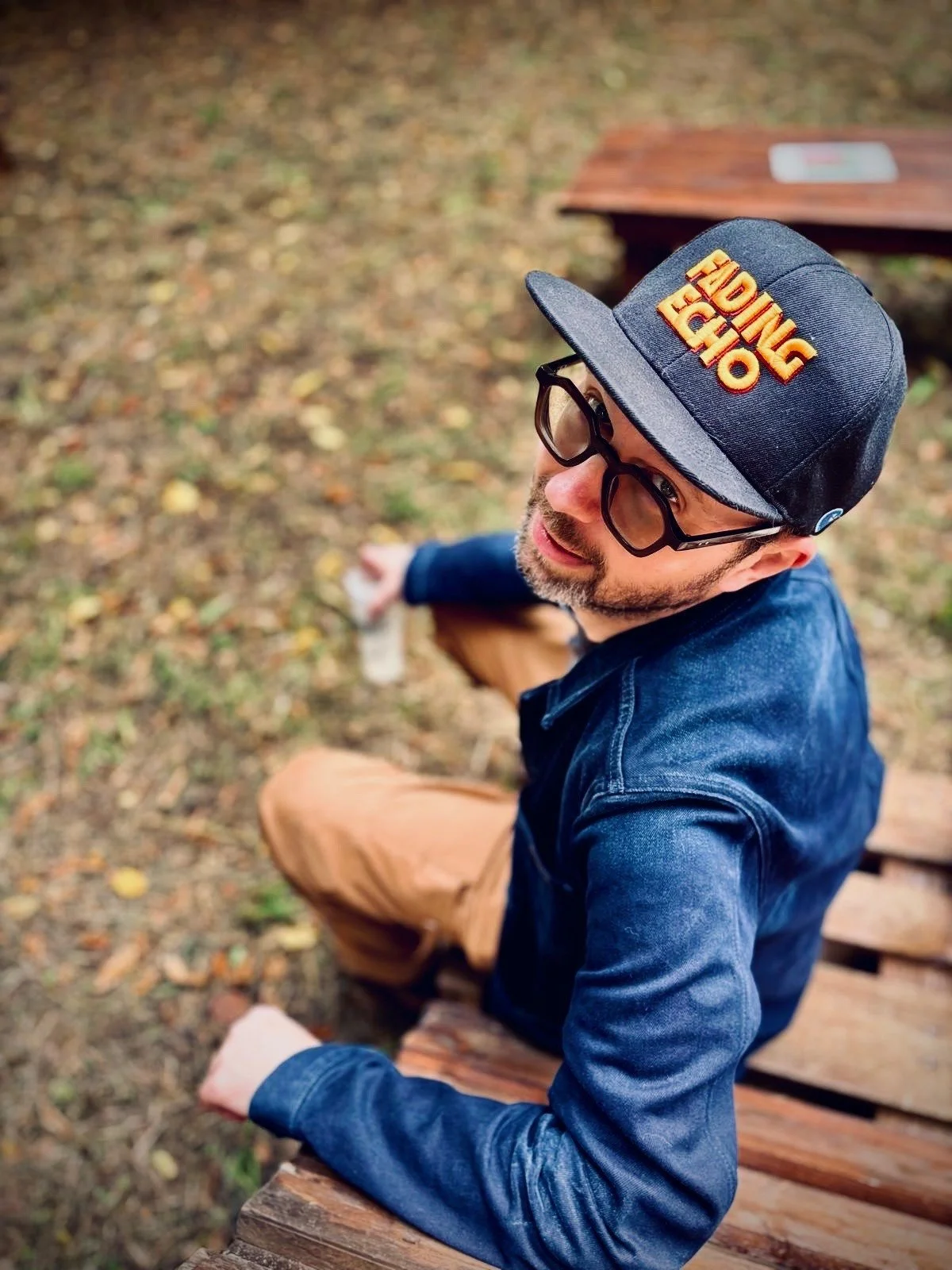Man sitting on a wooden bench outdoors in autumn, wearing glasses, a dark baseball cap with an orange logo, a denim jacket, and brown pants, looking up.