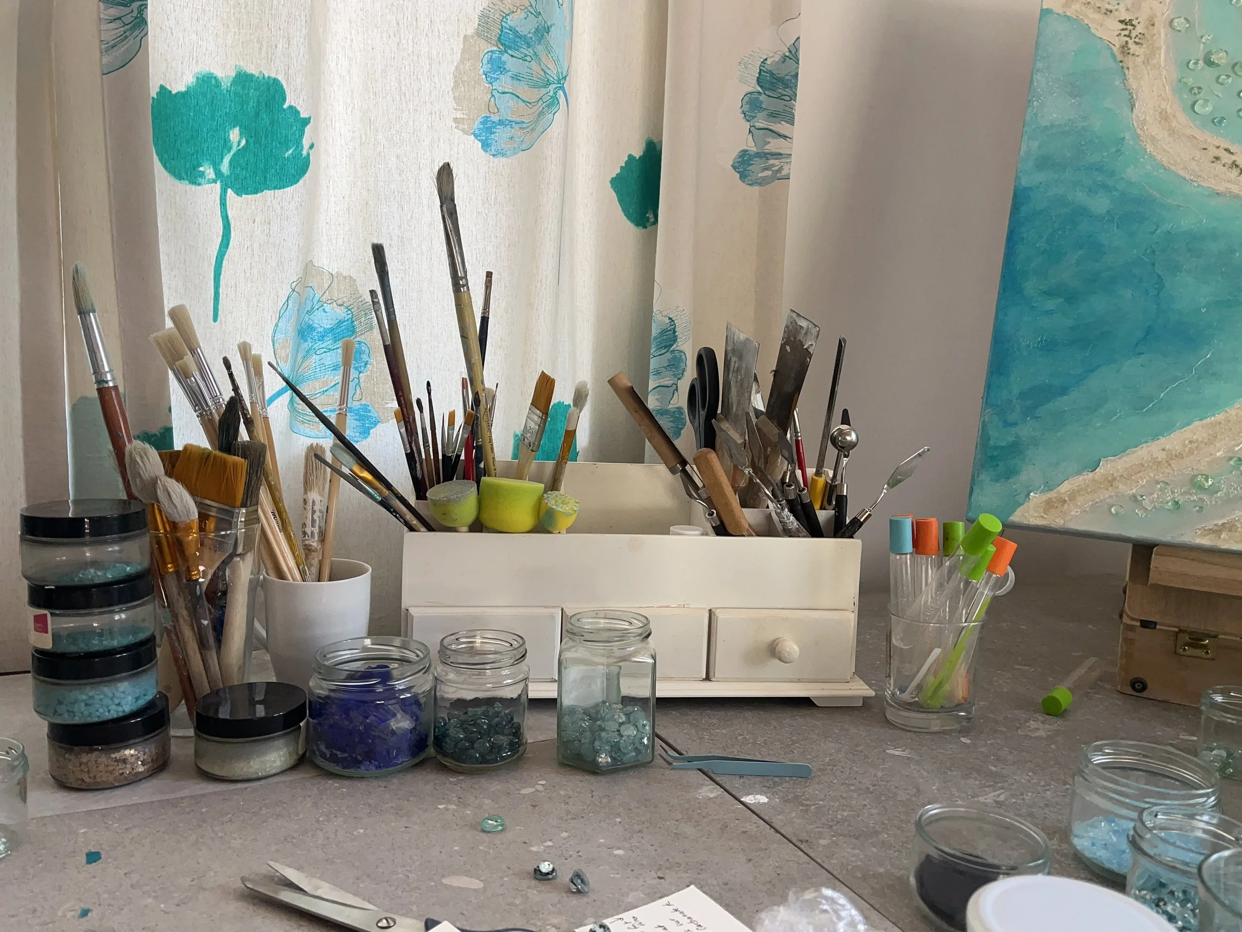 Un espace de travail artistique avec pinceaux, bocaux de peinture, et une peinture en cours représentant une scène balnéaire avec une plage et une mer turquoise.