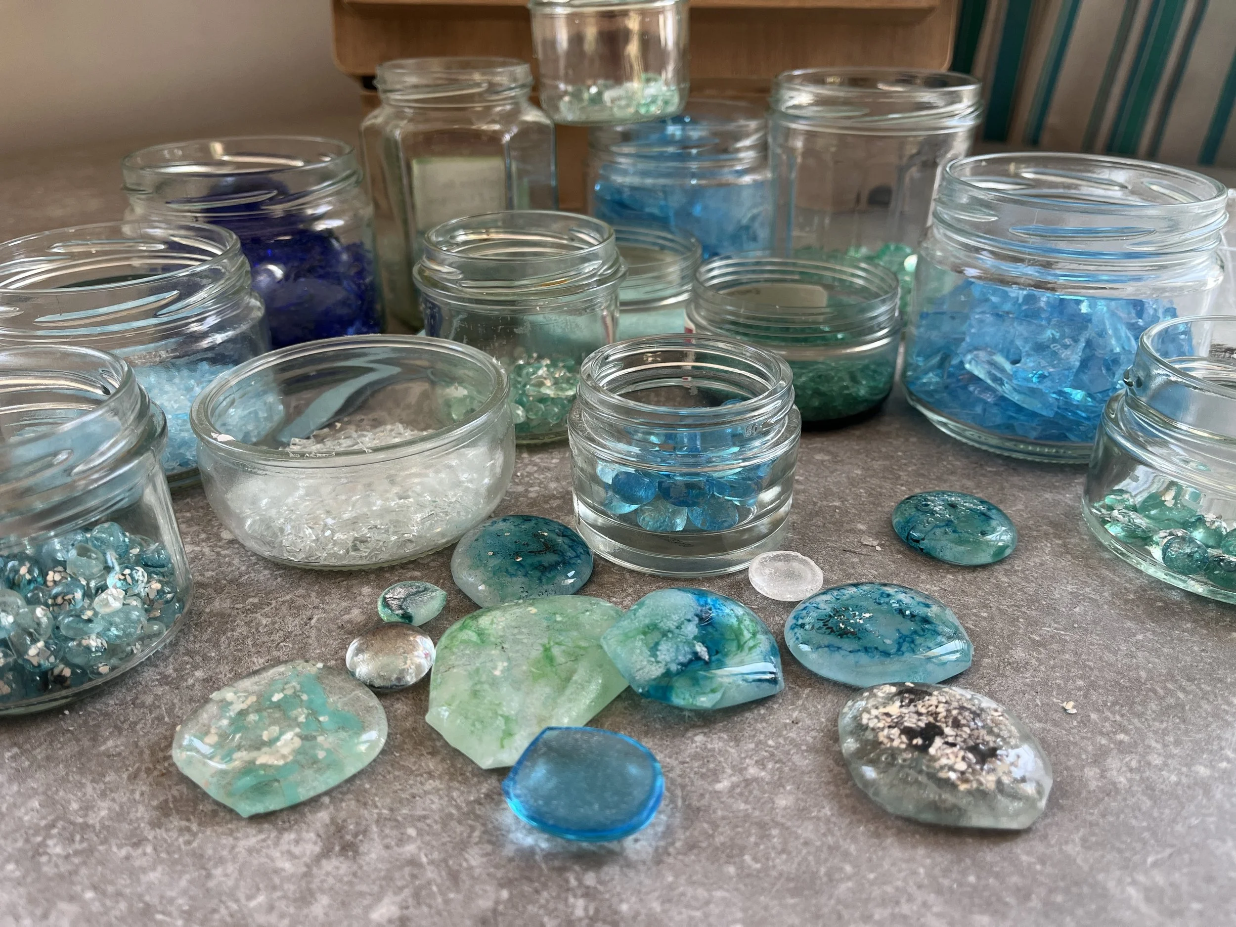 Différents bocaux en verre contenant des morceaux de verre de couleur bleu, vert, blanc, transparents et décorés, placés sur une surface en pierre ou en béton.