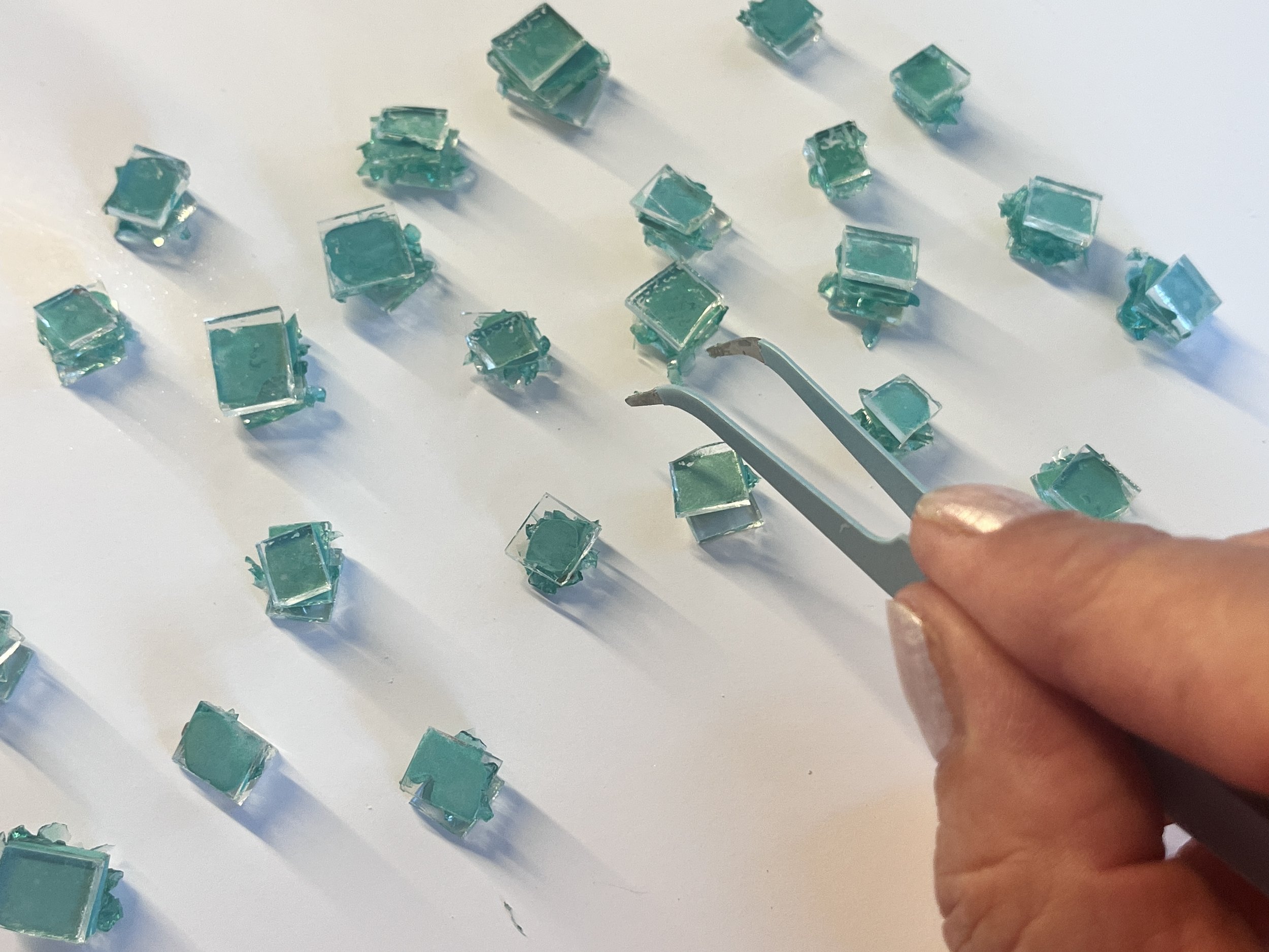 Une main tient un outil en métal. Autour, il y a plusieurs petits carrés en verre de couleur turquoise, distribués sur une surface blanche.