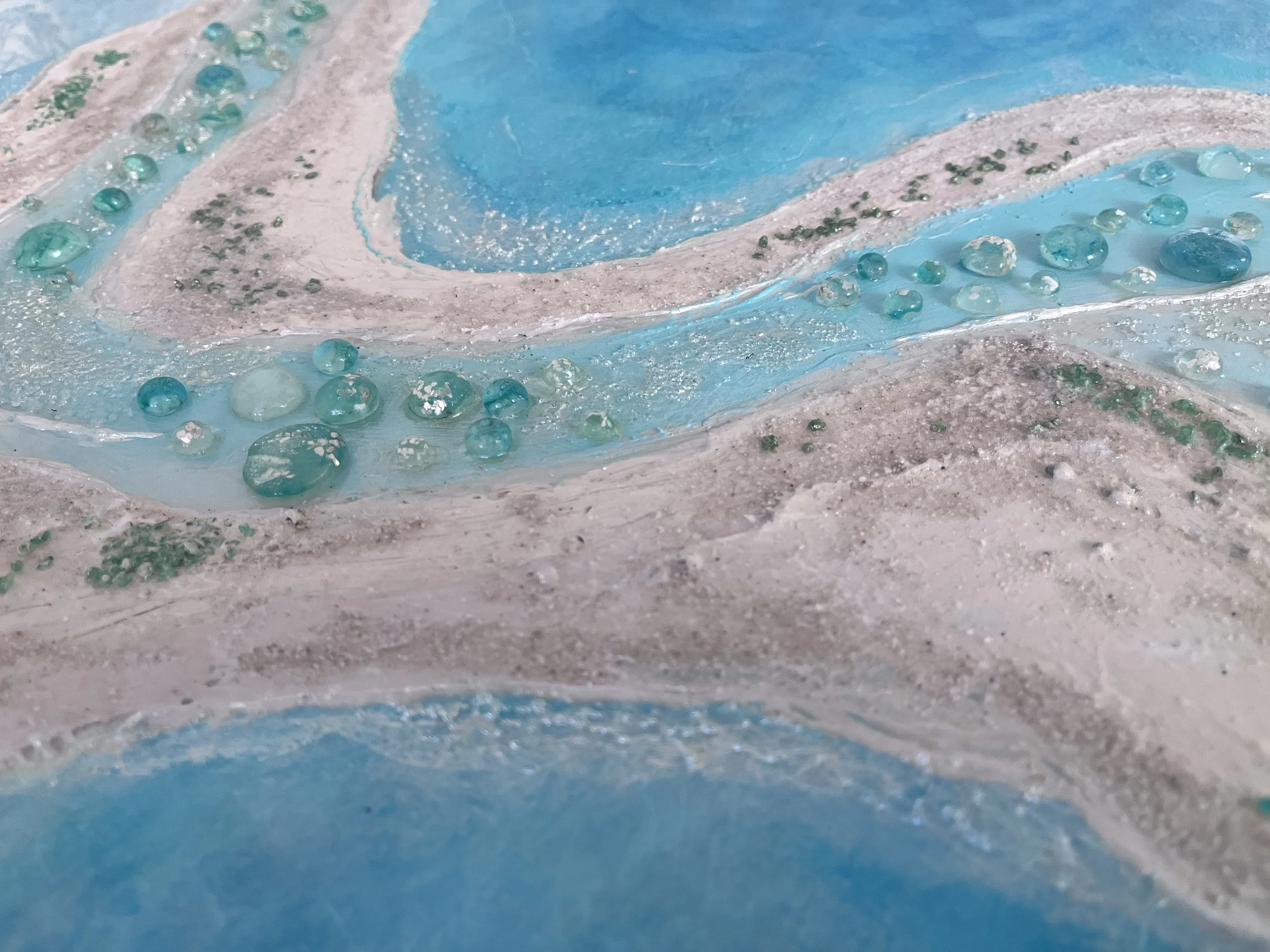 Surface brillante avec des perles de différentes tailles, principalement en teintes de bleu et de vert, sur une texture qui ressemble à du sable ou de la pierre, dans des tons pâles.
