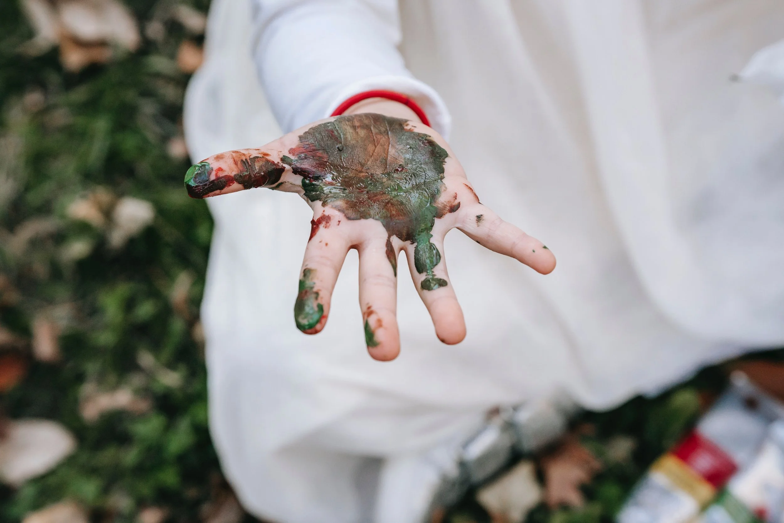 A child's hand covered in green, brown, and red paint, with some paint smeared on their fingers and palm, against a blurred background of greenery.