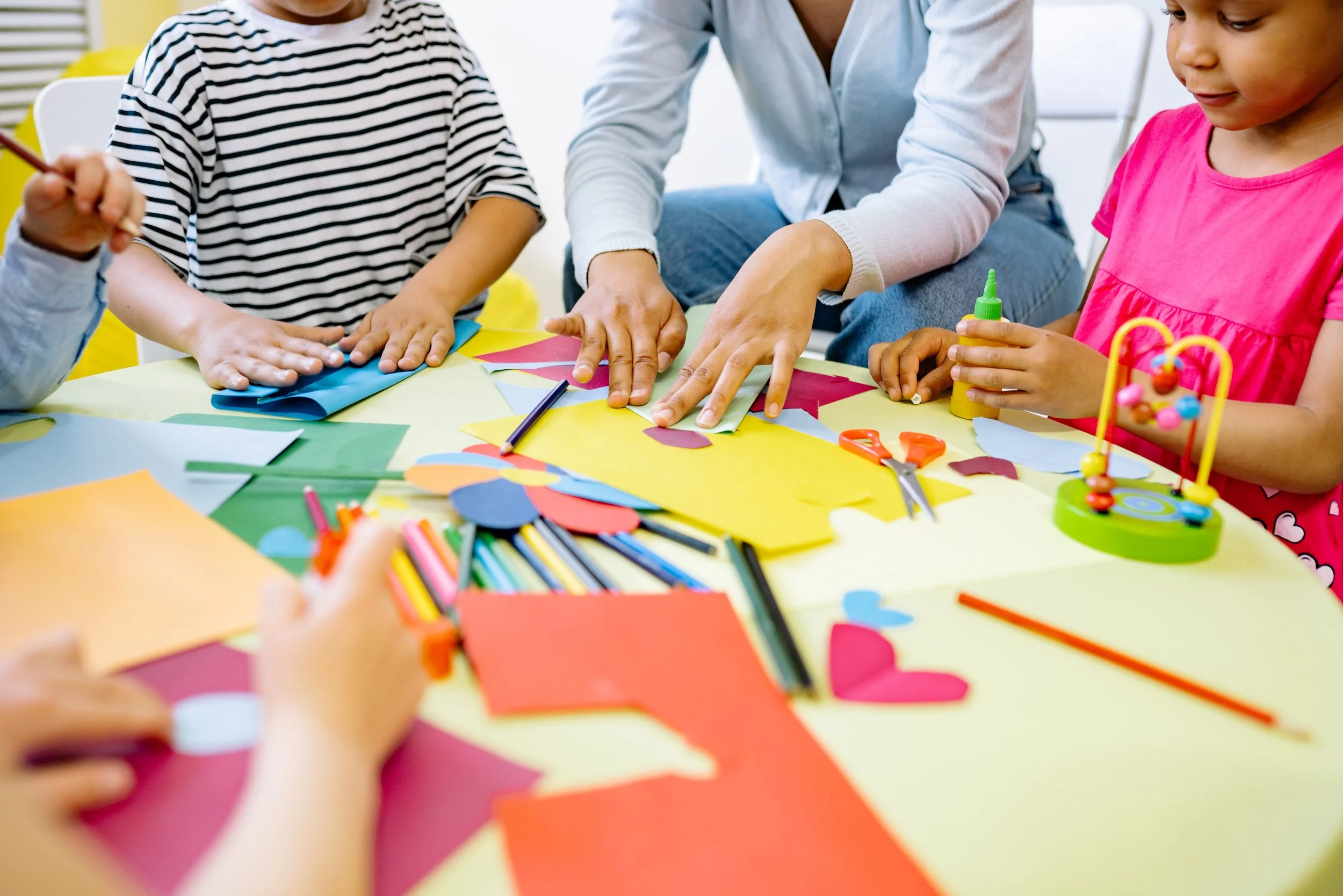Kids and an adult working together on arts and crafts at a table with colorful paper, scissors, glue, and markers.