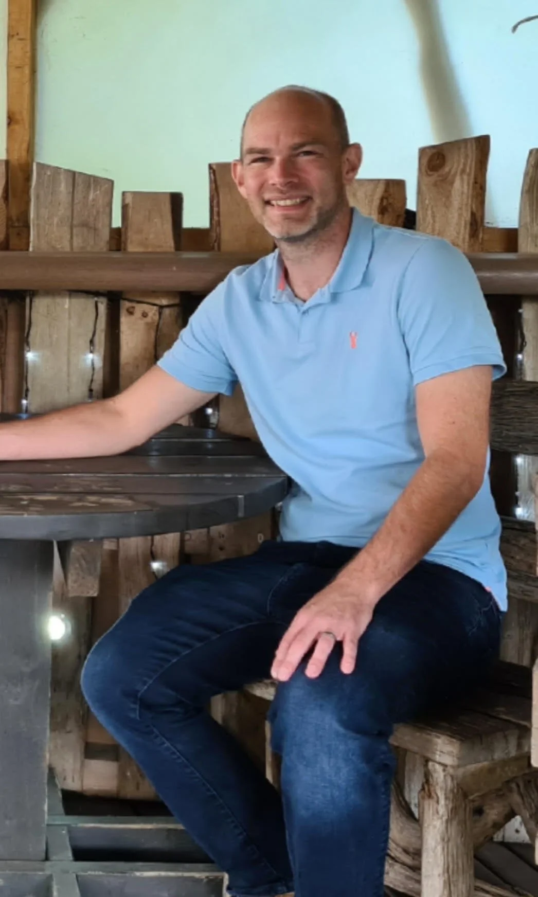 A man with short hair, wearing a light blue polo shirt and dark jeans, sitting at a rustic wooden table in a cozy setting with wooden decor and soft lighting.