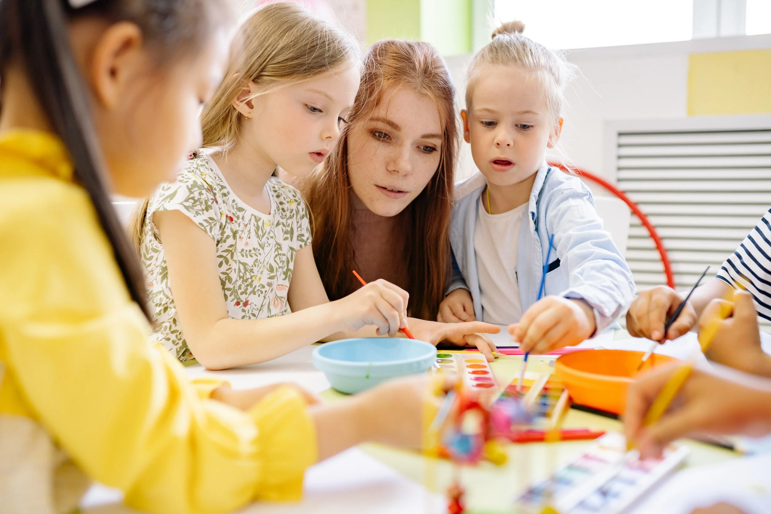 Children and an adult girl engaged in painting or craft activity at a table with paints and brushes.
