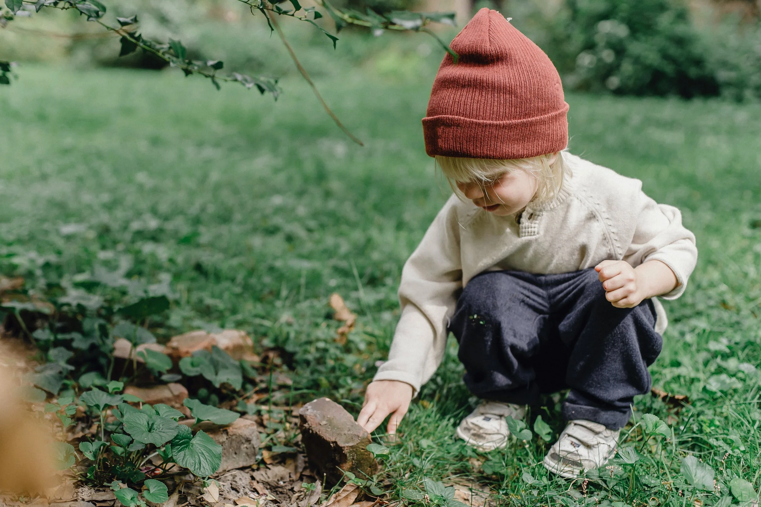 A young child with blonde hair wearing a red knit hat and beige jacket crouches down outdoors, exploring the grass and plants on the ground.